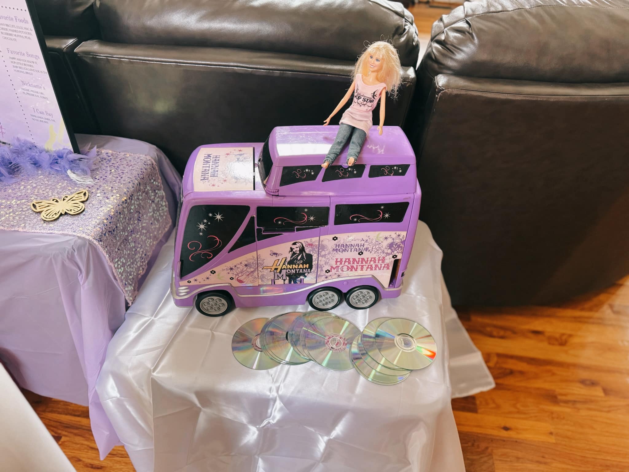 Hannah Montana tour bus toy with a doll sitting on top and several compact discs on the table below.