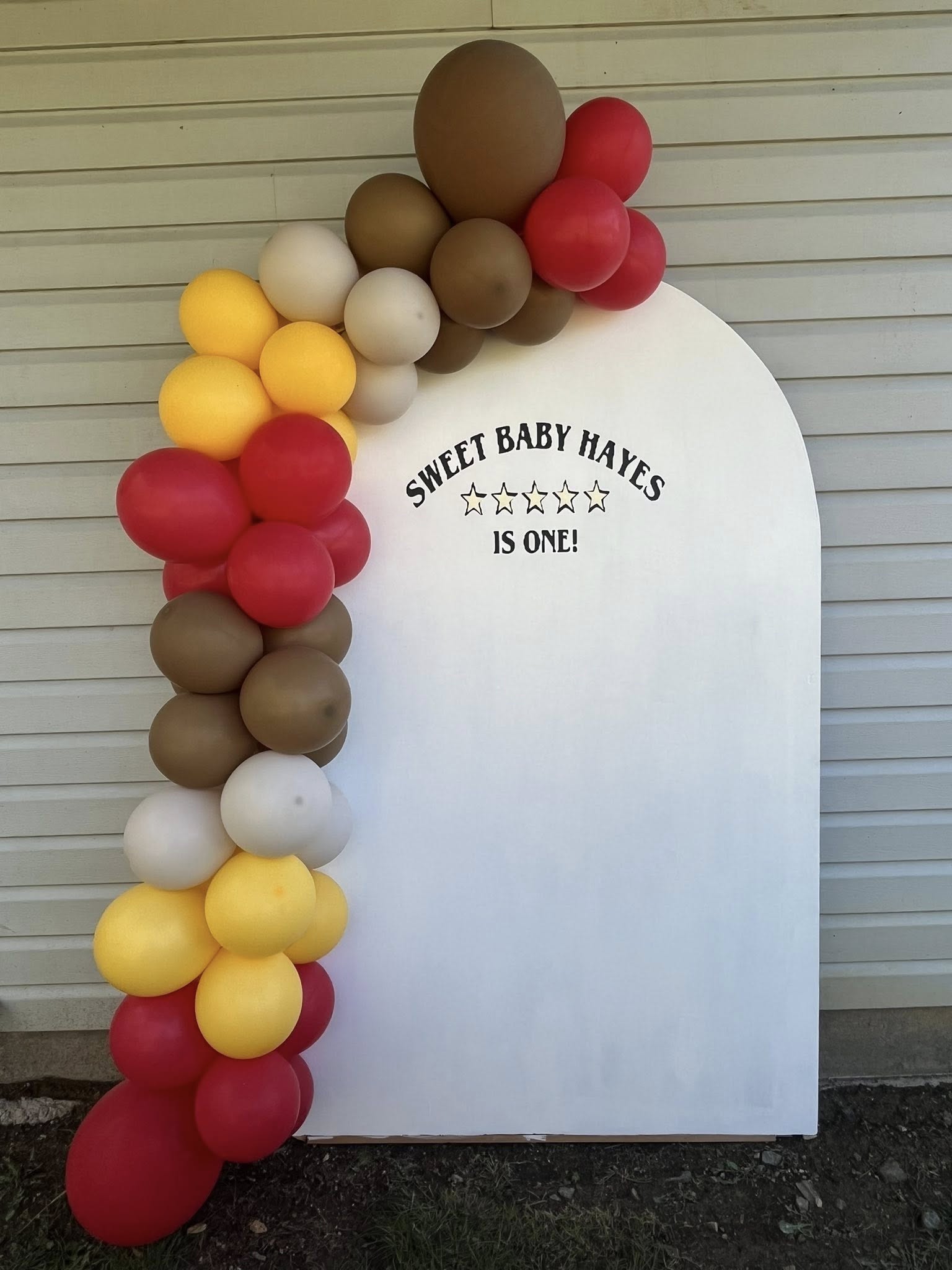 White arched backdrop with a balloon garland in yellow, red, brown, and white, saying Sweet Baby Hayes Is One.