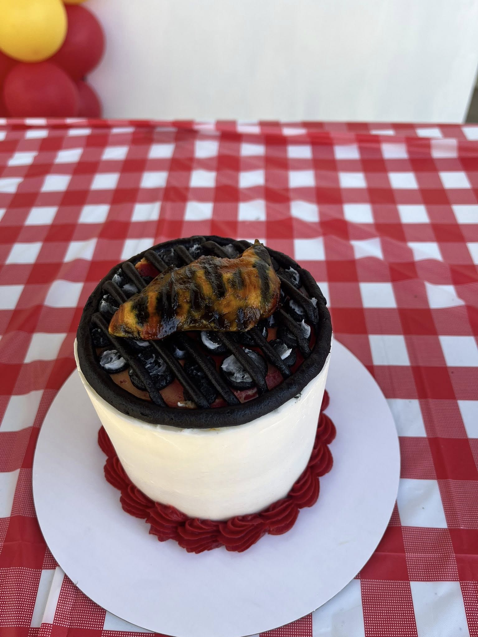 Round cake decorated to look like a small charcoal grill with a grilled chicken piece on top, sitting on a red and white checkered tablecloth.