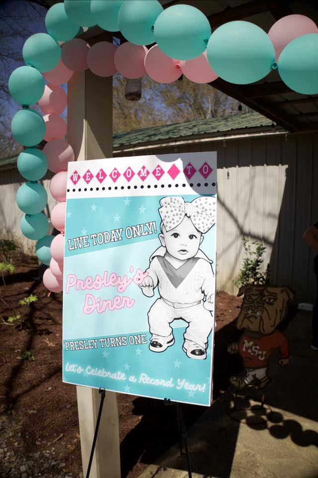 Welcome sign for Presley's first birthday party with a diner theme, decorated with a teal and pink balloon arch.