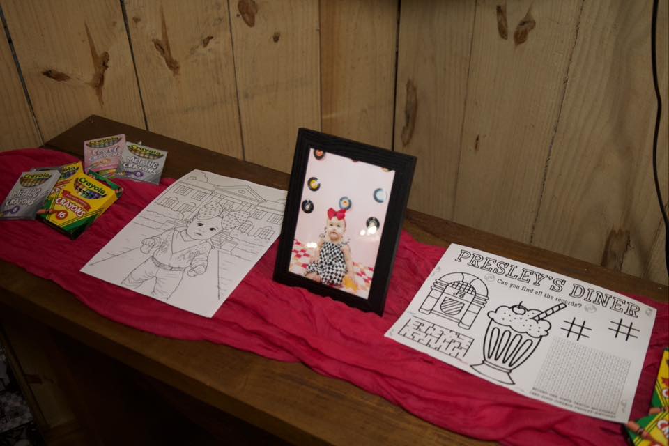 Activities table with crayons, coloring pages, and a framed photo of a baby dressed like Elvis at a party.