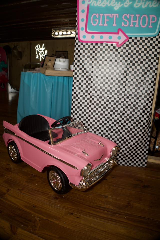 Vintage-style pink toy pedal car positioned in front of a checkered backdrop labeled Presley's Diner Gift Shop.