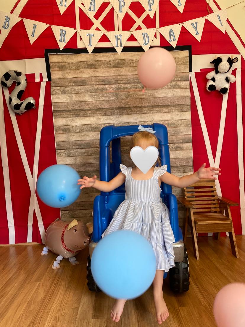 Toddler in a blue sundress celebrating a birthday in front of a barn-themed backdrop with balloons.