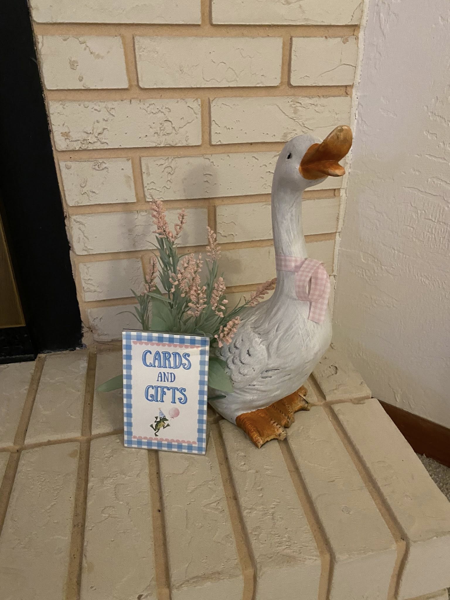 Ceramic decorative goose wearing a pink gingham scarf next to a Cards and Gifts sign with a party frog illustration.