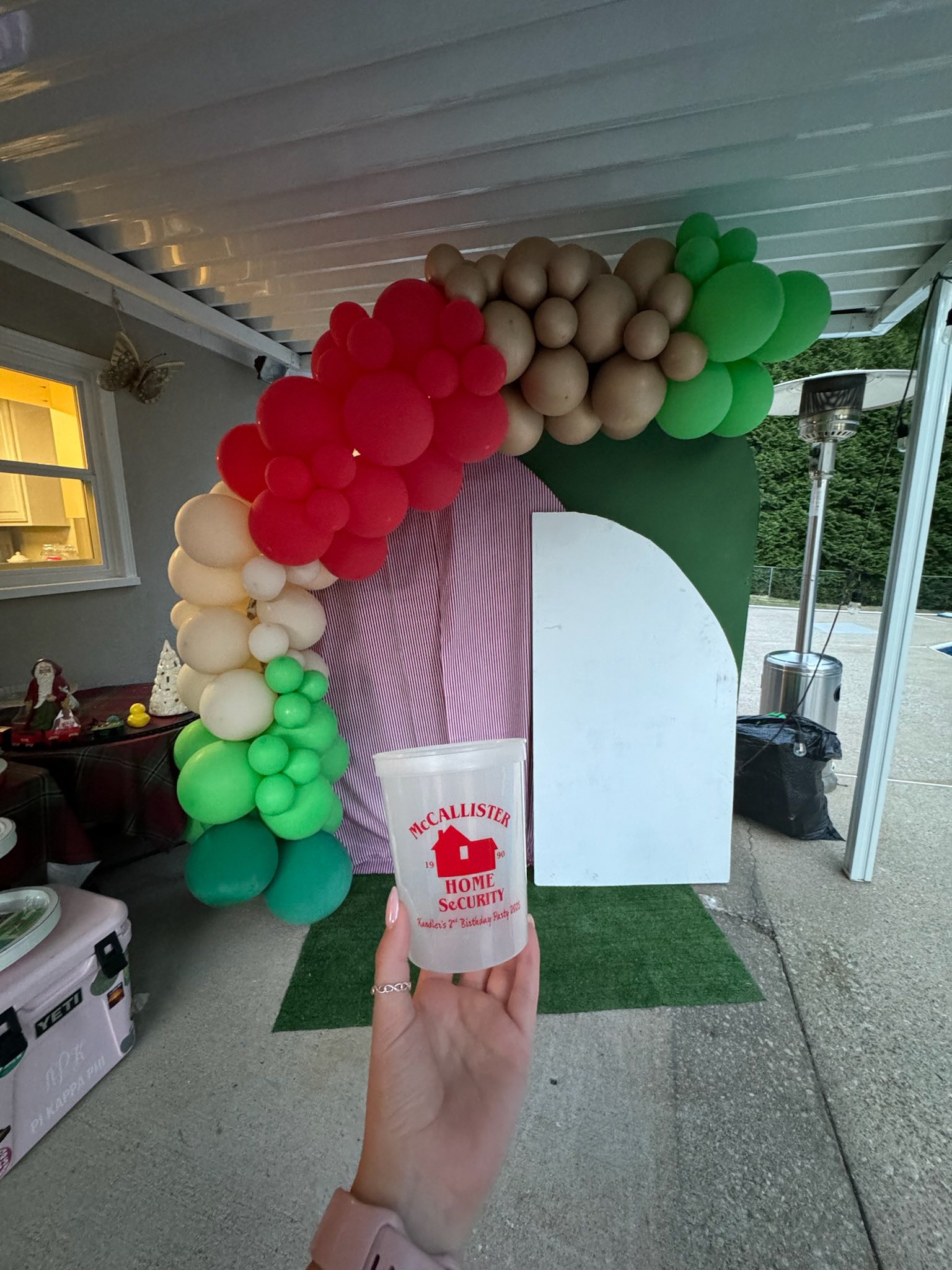 Custom plastic party cup featuring red McCallister Home Security branding in front of a holiday balloon arch.
