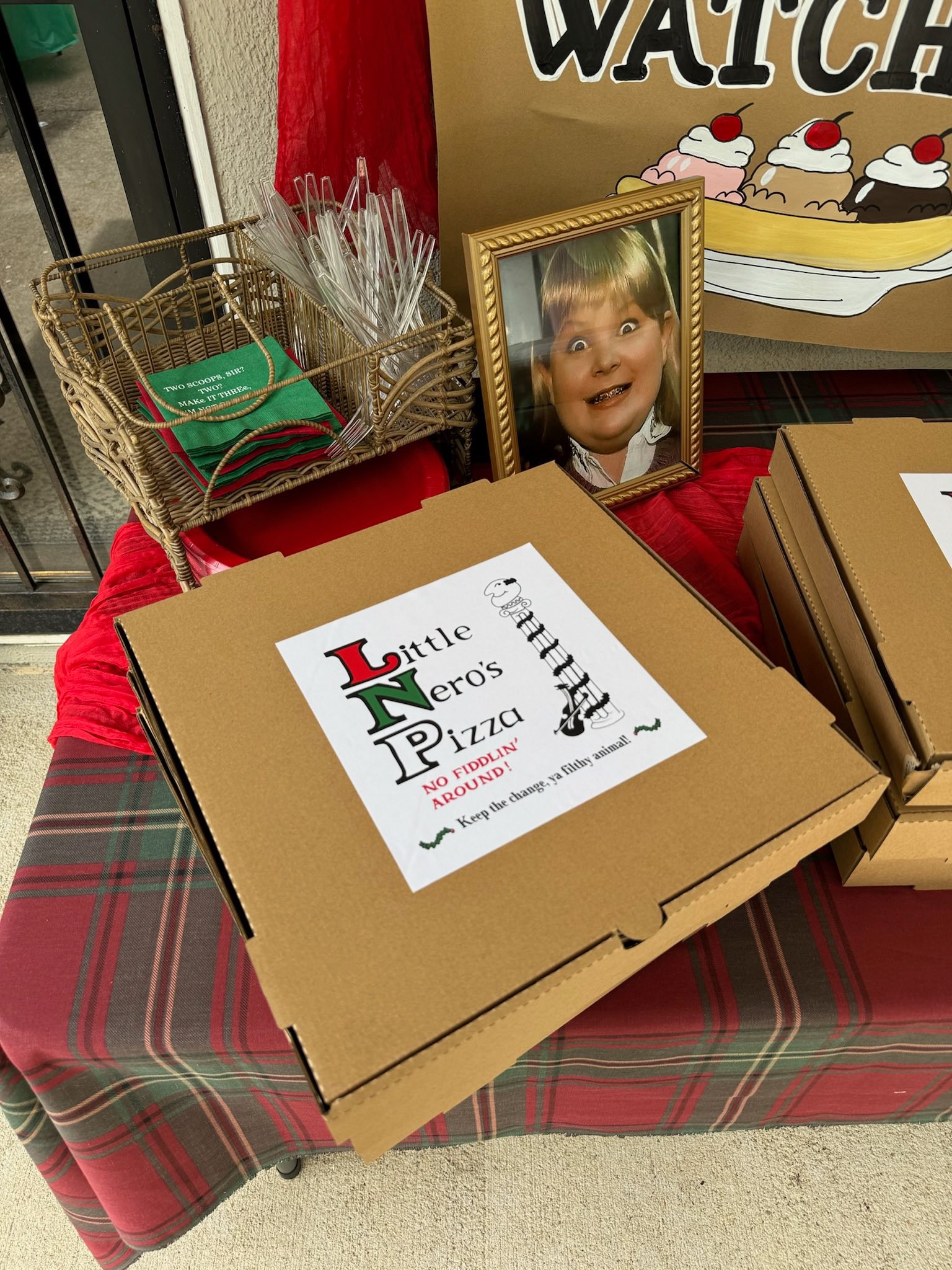 A cardboard pizza box with a custom Little Nero's Pizza sticker on a plaid table cloth with home-themed party decor.