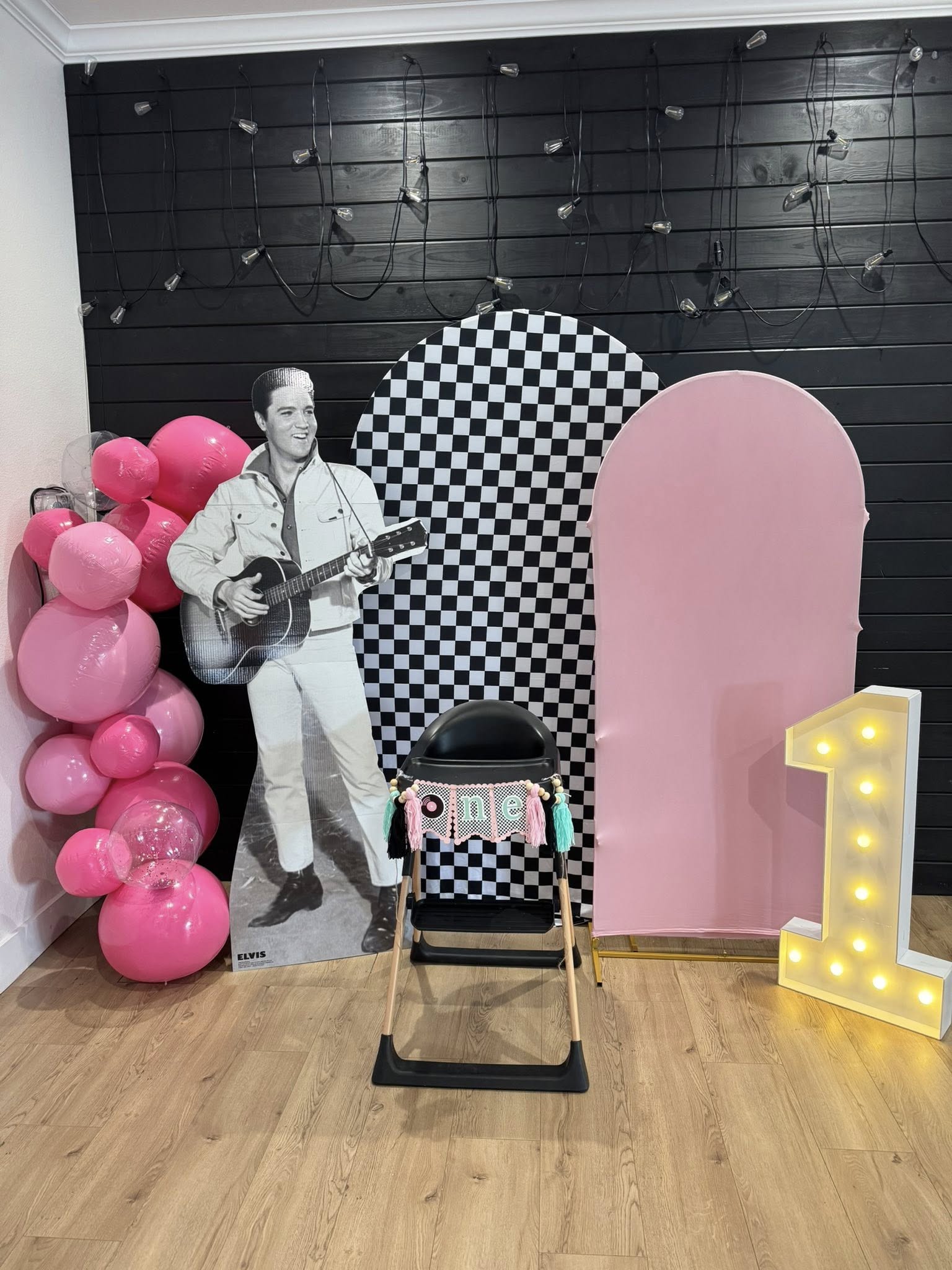 Elvis-themed first birthday party setup with high chair, checkered backdrop, balloons, and light-up number one.