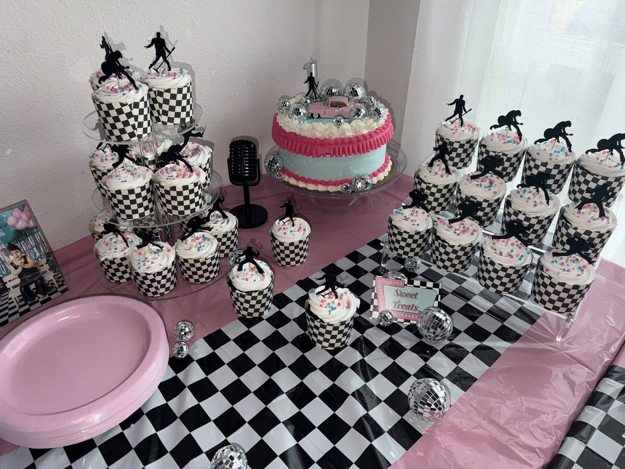 1950s rock and roll themed party table with a pink and teal cake and cupcakes featuring checkered wrappers.