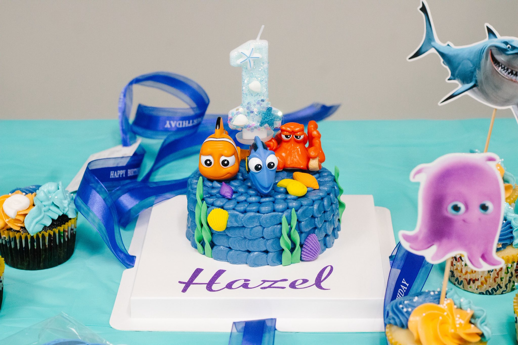 Finding Nemo themed first birthday cake with fondant sea creature decorations and a number one candle on a blue table.