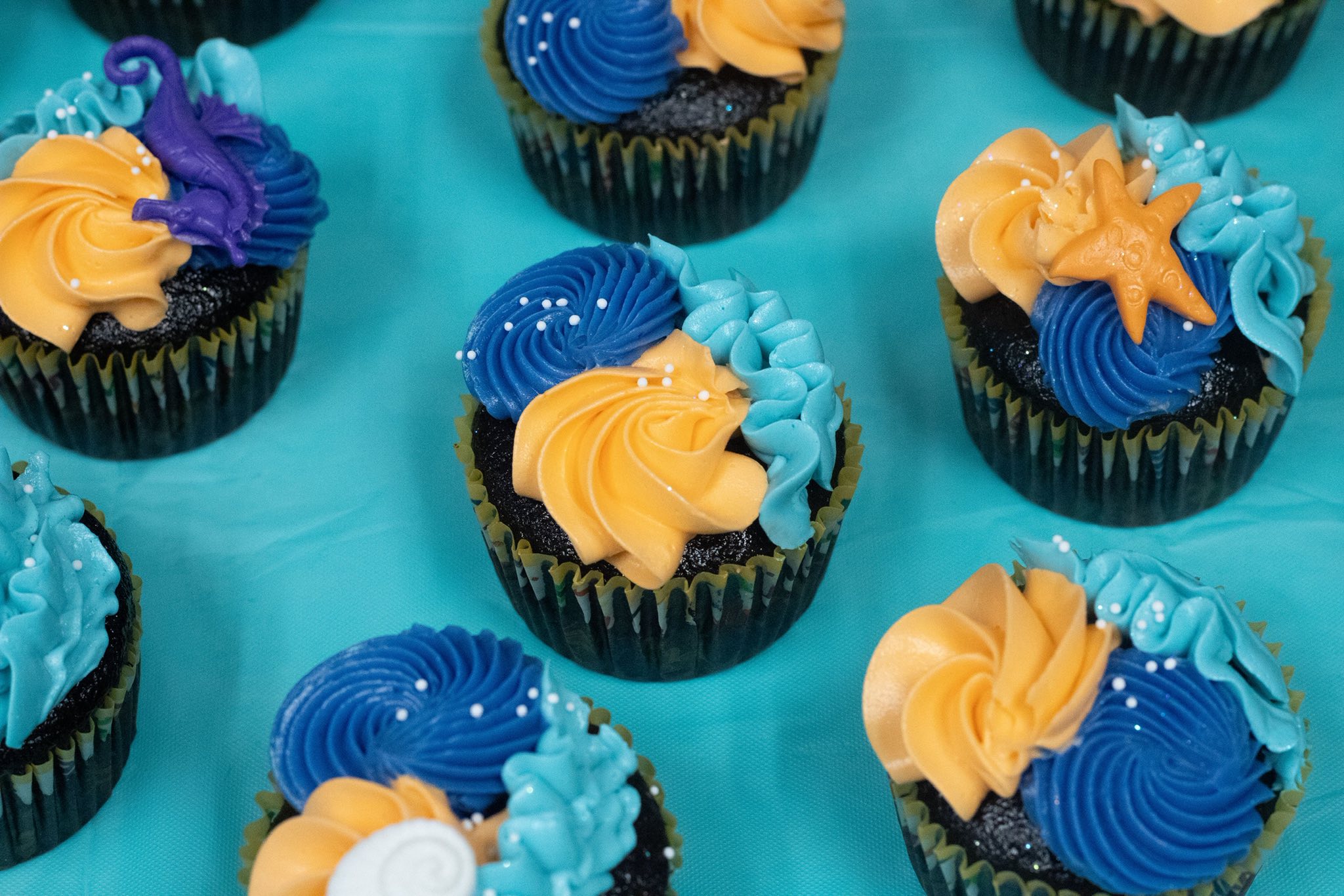 Assorted cupcakes decorated with blue, orange, and light blue frosting swirls and nautical themed fondant shapes.
