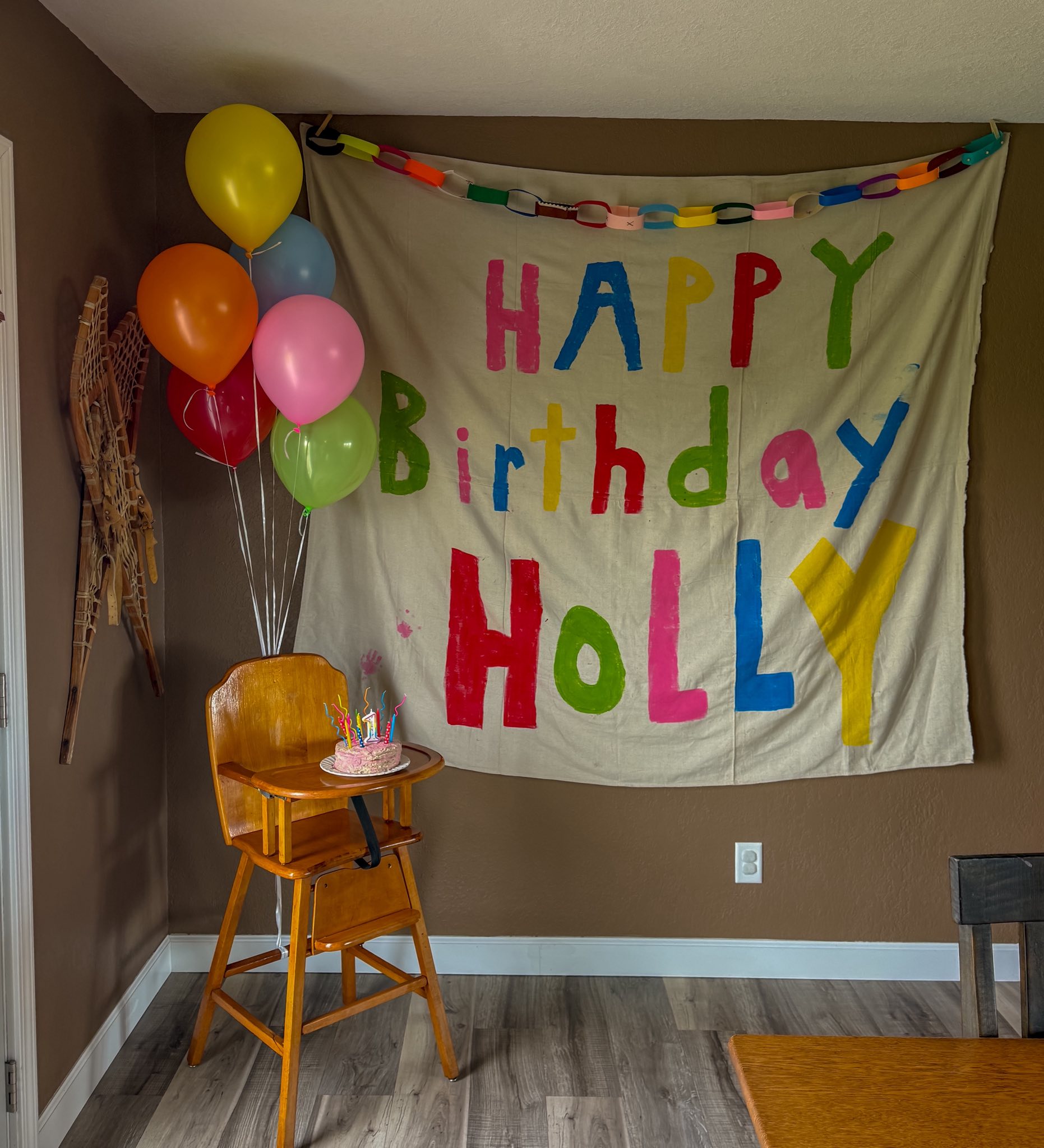 A first birthday setup featuring a small cake on a wooden high chair with a Happy Birthday Holly banner and colorful balloons.