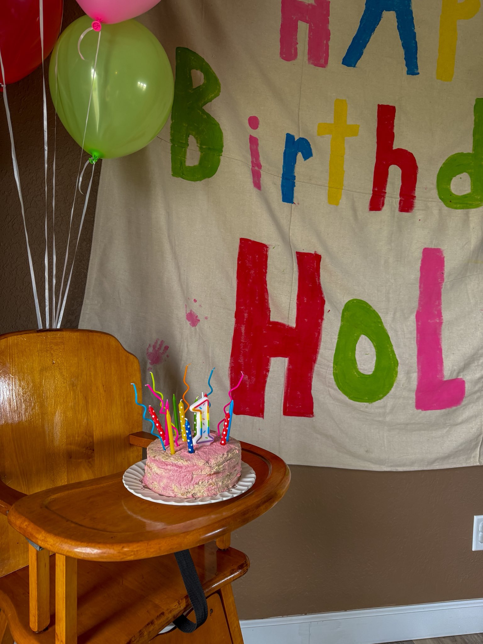First birthday cake with colorful candles on a wooden high chair in front of a painted birthday banner.