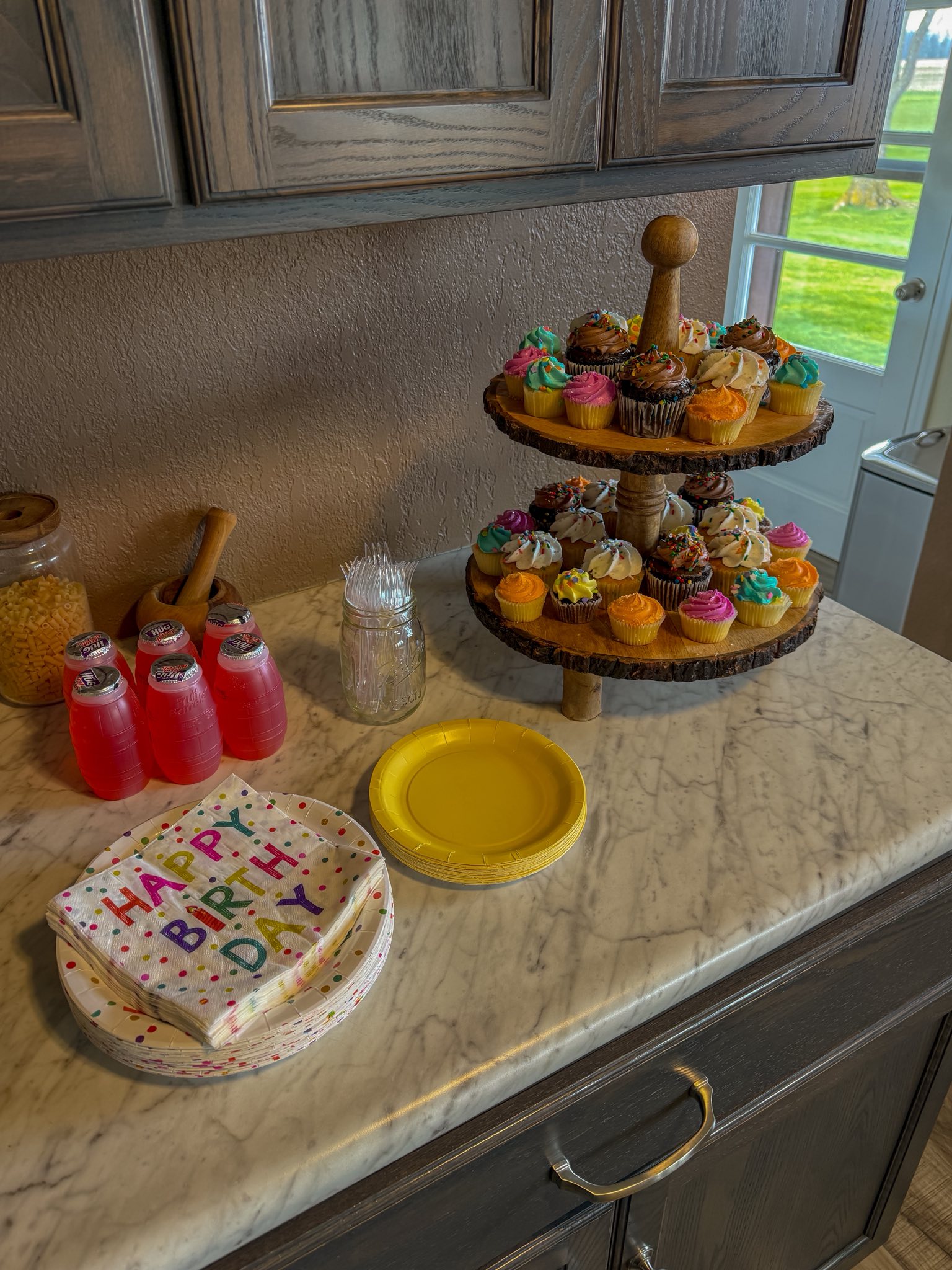 Two-tiered wooden stand filled with colorful cupcakes, served with juice bottles, napkins, and plates for a party.