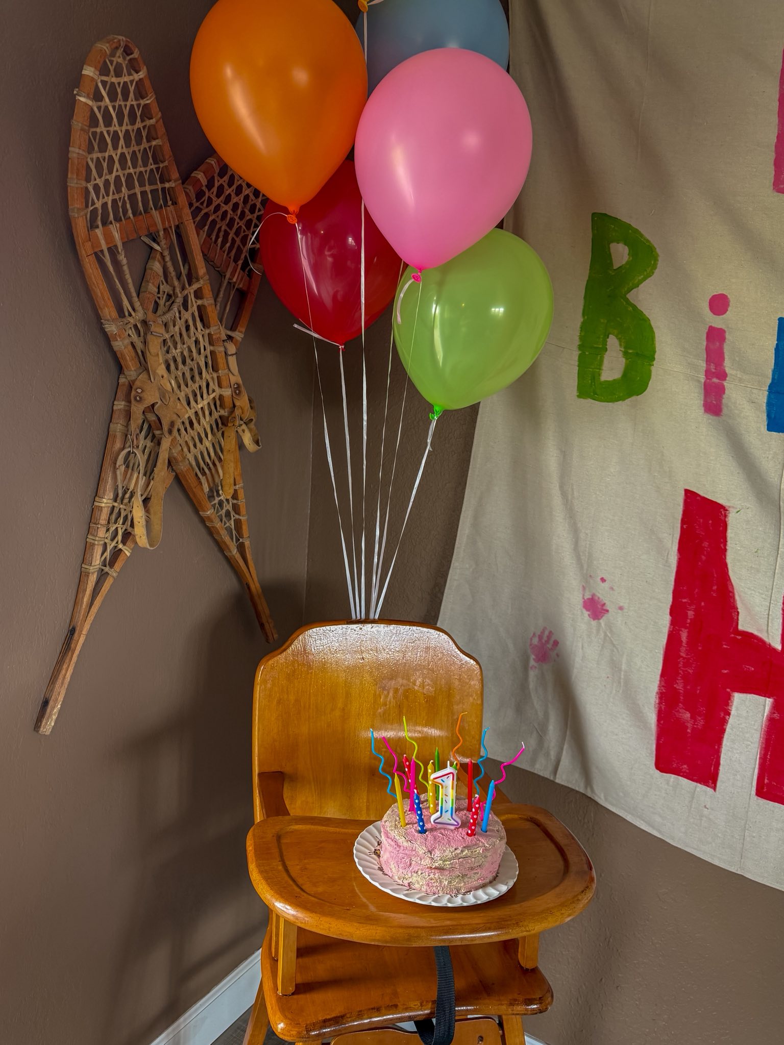 First birthday cake with colorful candles on a wooden high chair, framed by balloons and birthday decorations.