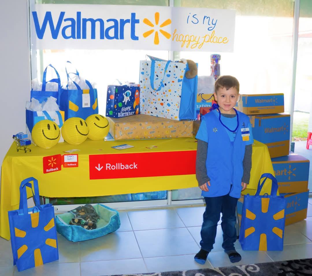 Young boy wearing a blue Walmart associate vest standing in front of a party table with gifts and shopping bags.