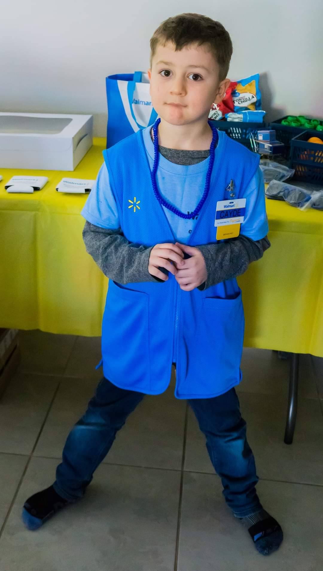 Young child dressed in a Walmart employee uniform vest and name tag standing in front of a yellow party table.