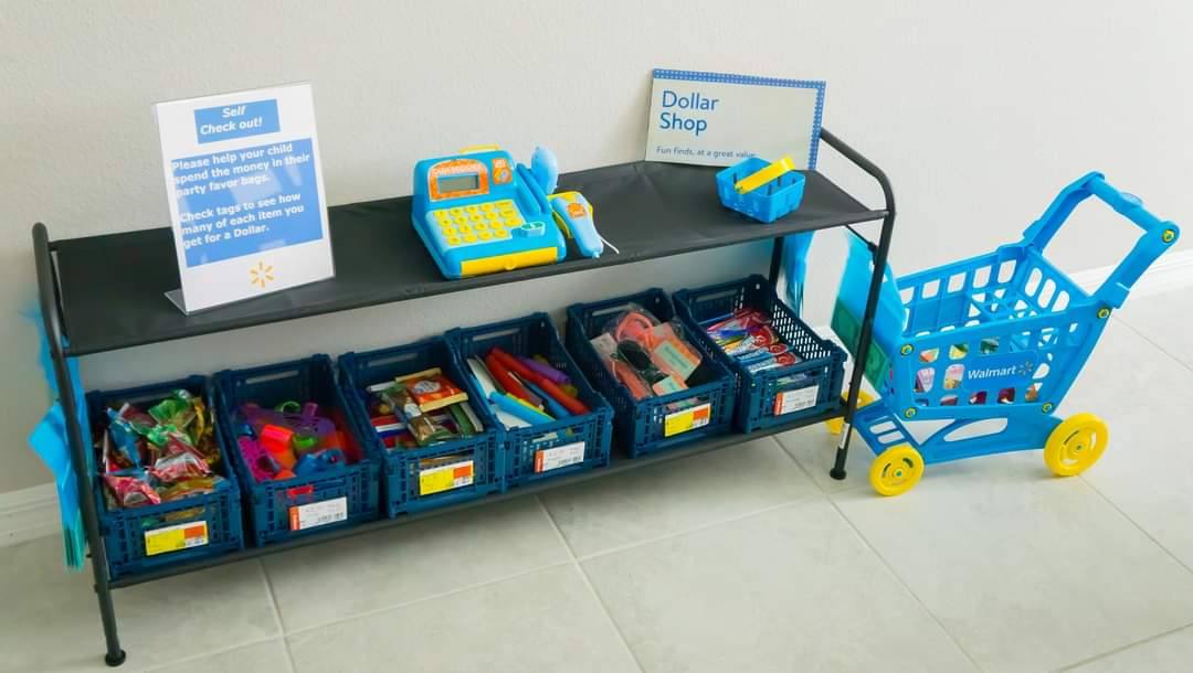 Toy dollar shop setup with bins of party favors, a toy cash register, and a blue toy shopping cart.