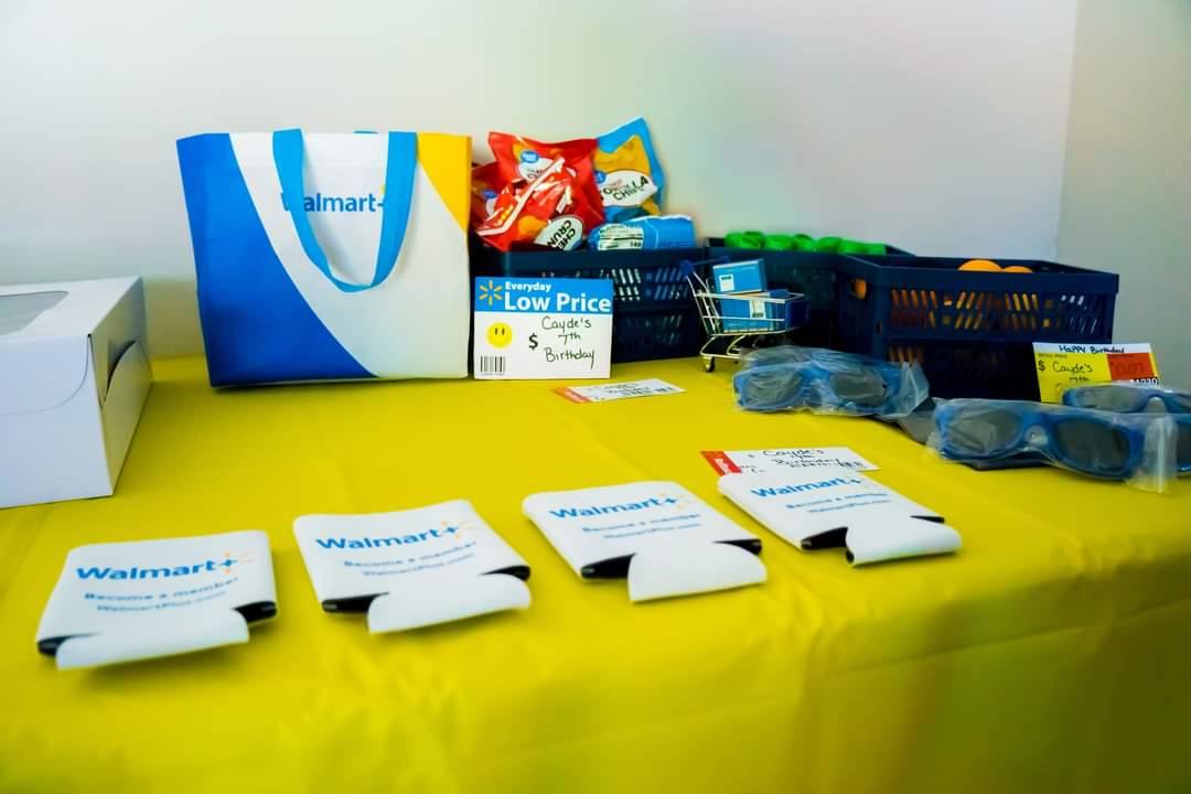 Walmart+ branded party favors, snacks, and decorations arranged on a yellow tablecloth for a birthday celebration.