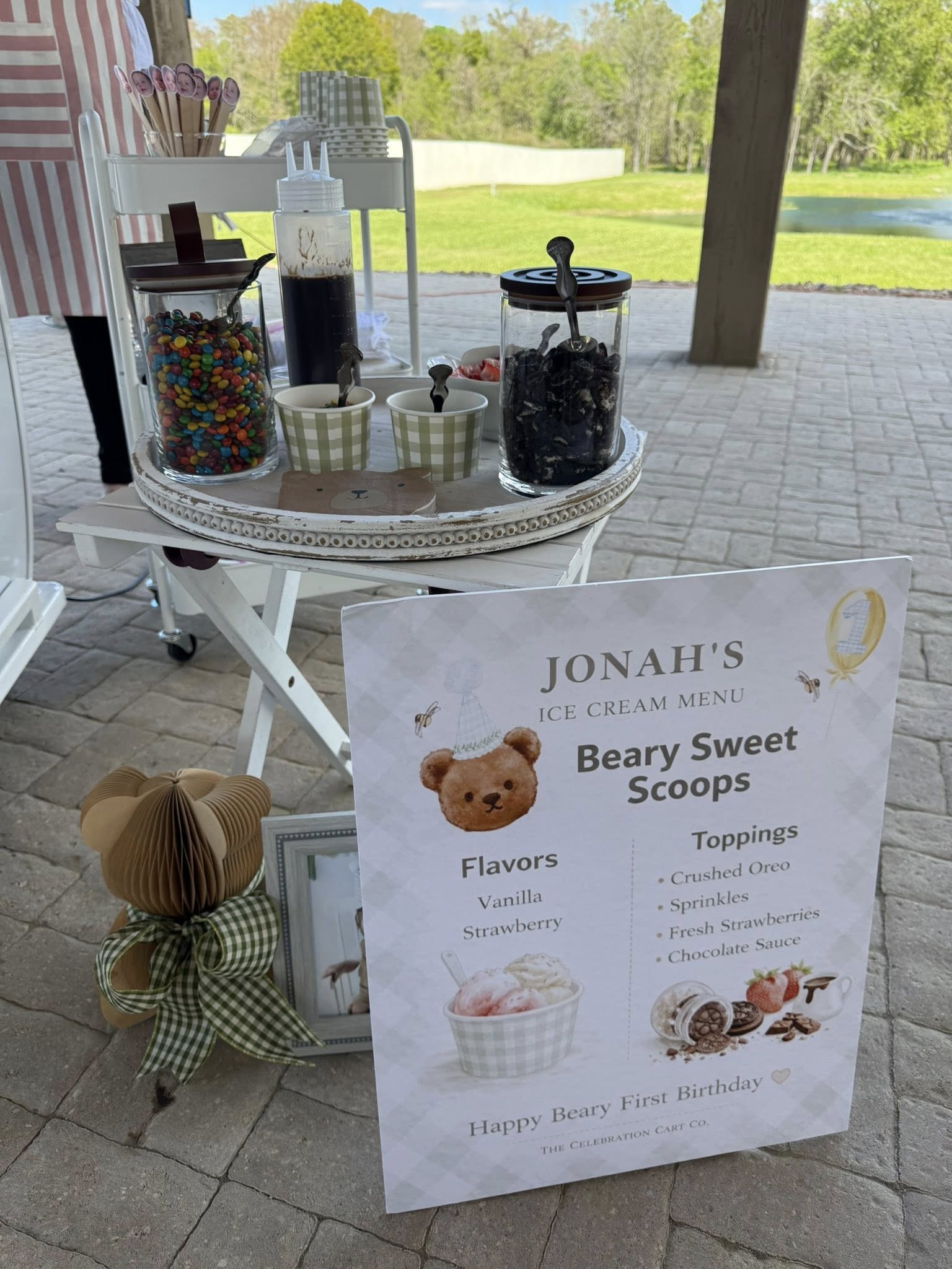 Bear-themed first birthday ice cream cart menu sign with matching jars of toppings including candy and chocolate.