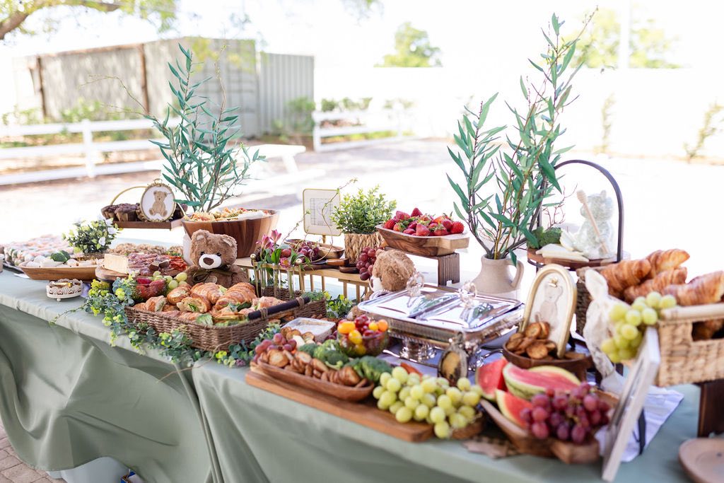 Table spread for a party featuring a charcuterie board, fresh fruit, croissants, and teddy bear decorations.