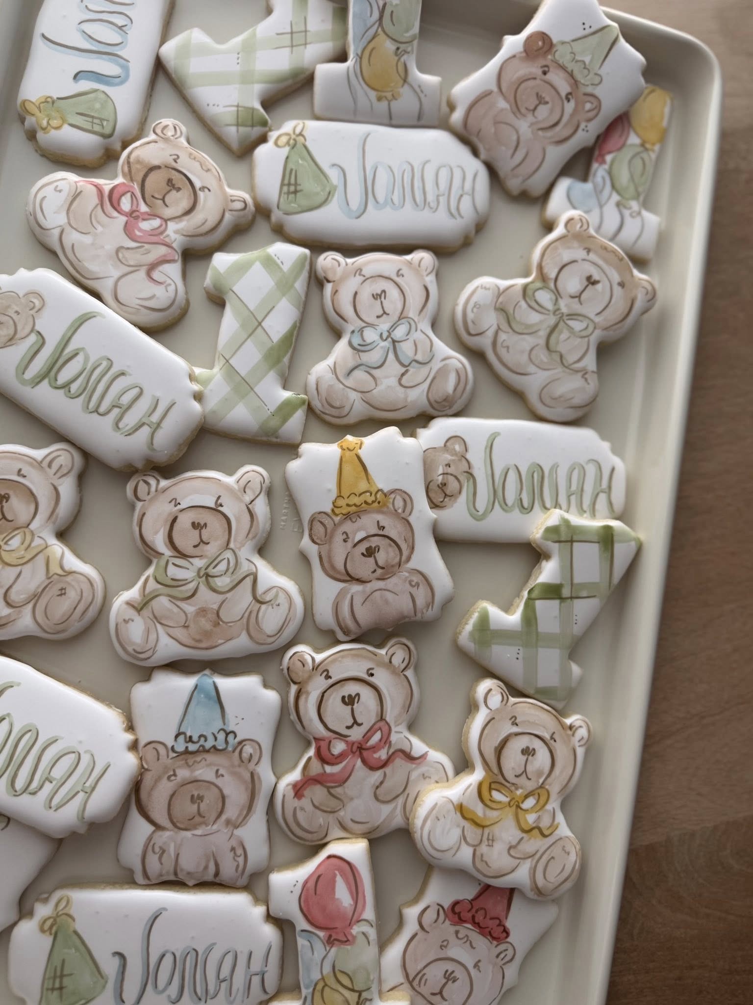 Assorted decorated sugar cookies for a first birthday, including watercolor-style teddy bears, plaques with the name Jonah, and number-one shapes.