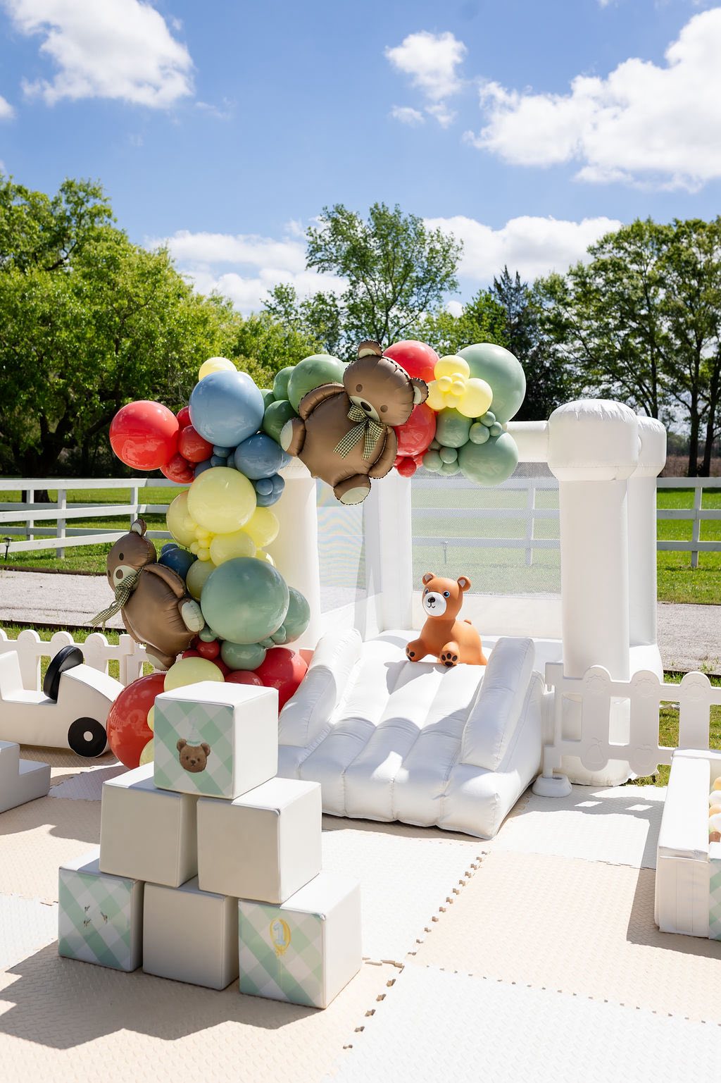 White inflatable bounce house with bear-themed balloon arch, soft play blocks, and a toy bear outdoors.