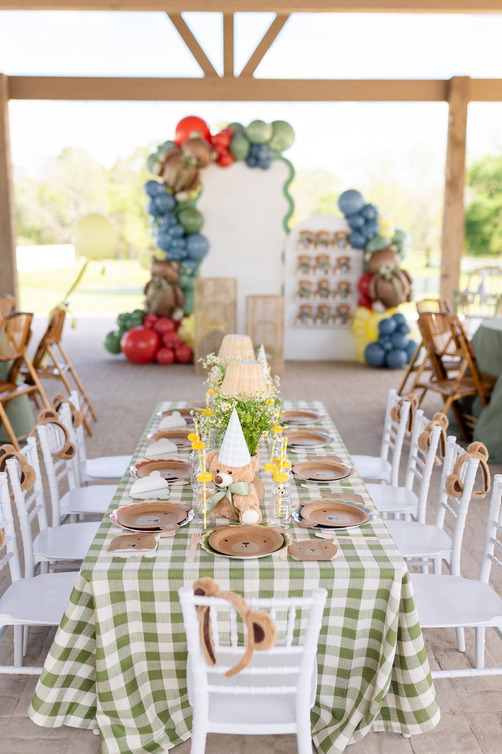 Teddy bear themed party table setup with green plaid tablecloth, bear shaped plates, and balloon arch backdrop.