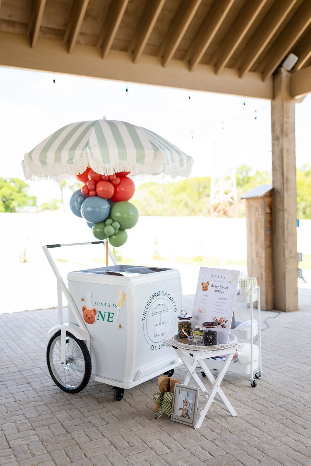 A white ice cream cart with a green striped umbrella and teddy bear first birthday decorations.