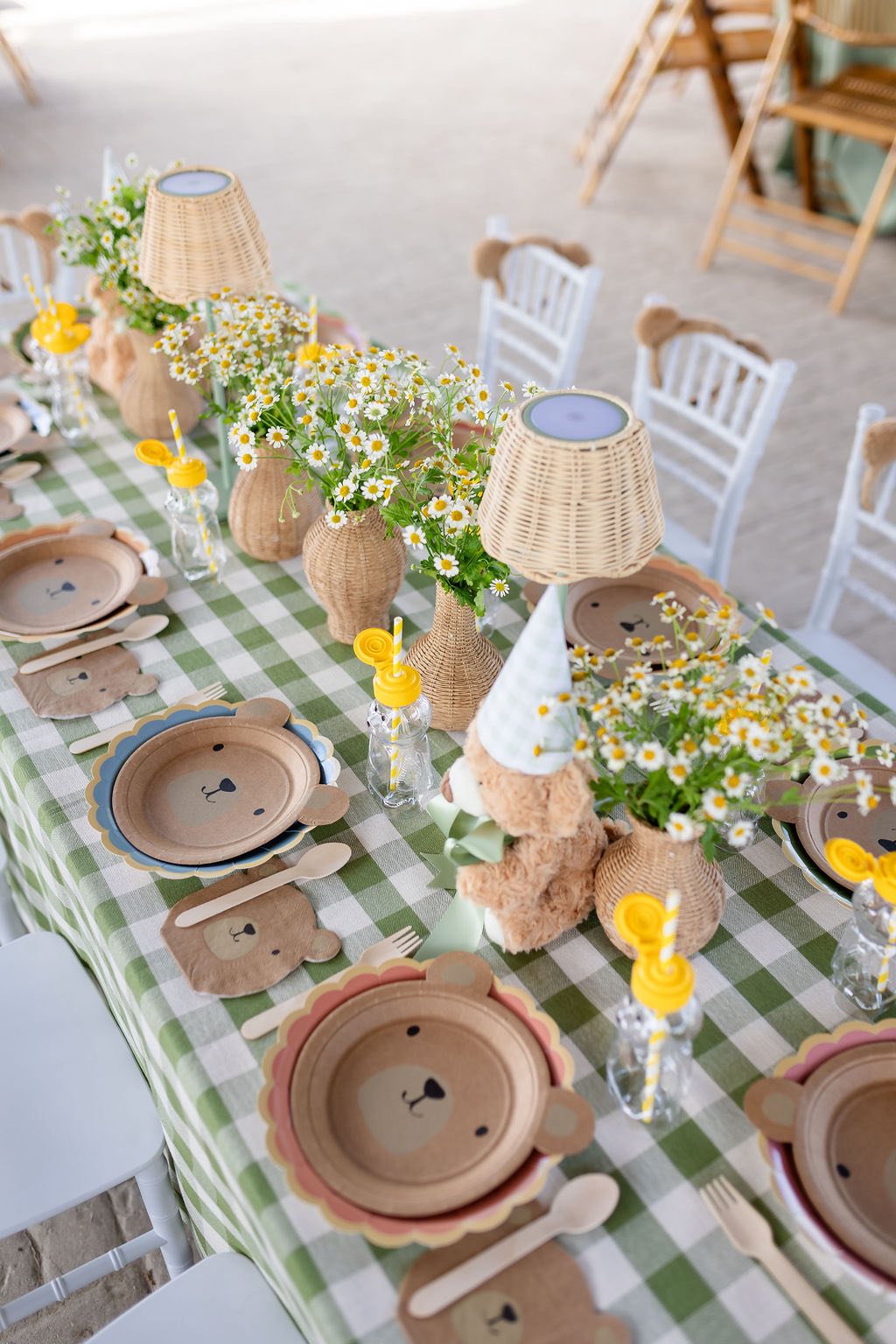 Table set for a bear-themed party with gingham tablecloth, bear-faced paper plates, and daisy centerpieces.