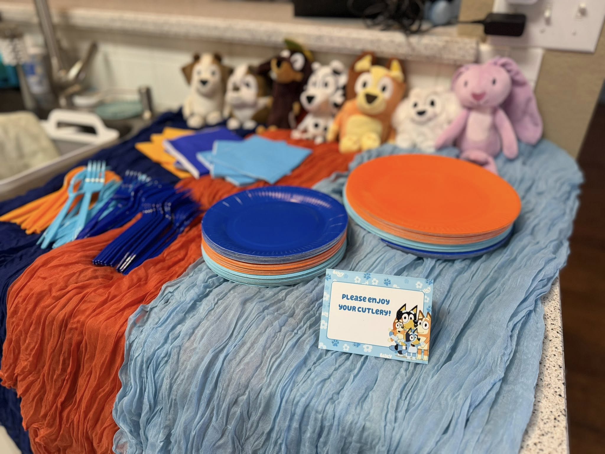 Bluey-themed party table set up with orange and blue plates, cutlery, napkins, and plush toy characters.