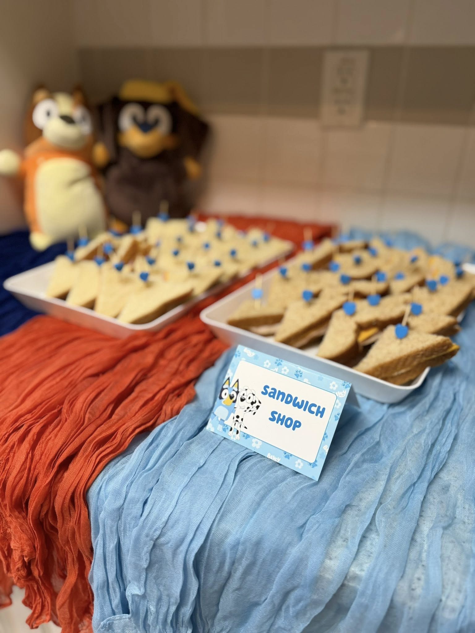 Platters of triangle sandwiches with blue heart picks at a Bluey-themed party with a Sandwich Shop sign.
