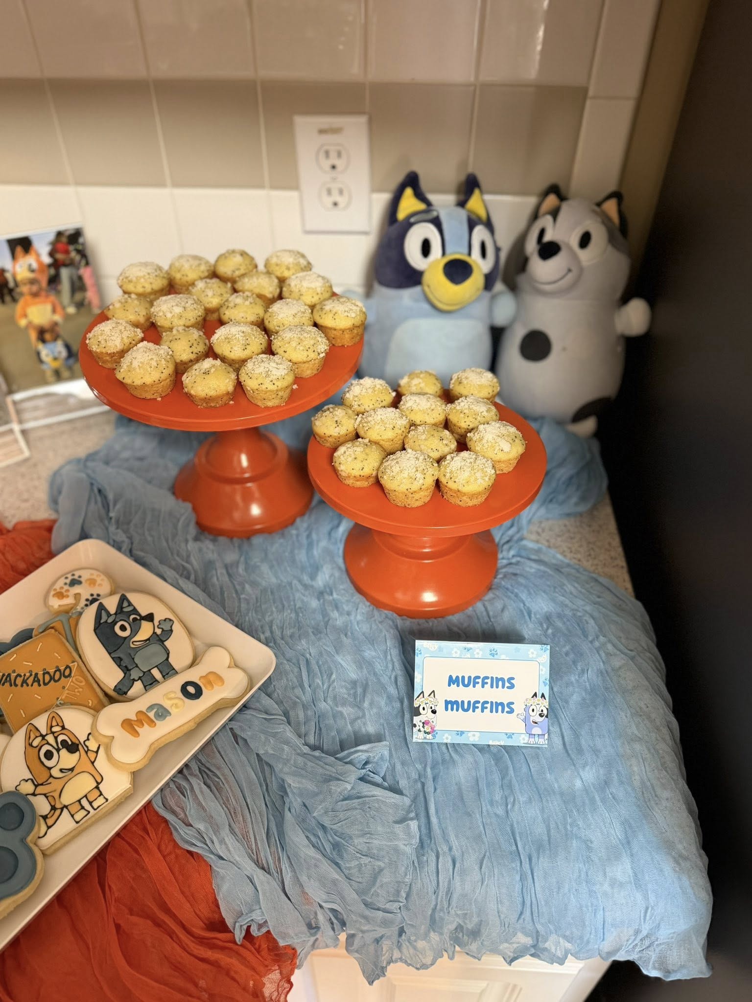 Bluey-themed party display with poppy seed muffins on orange cake stands, custom cookies, and Bluey plush toys.
