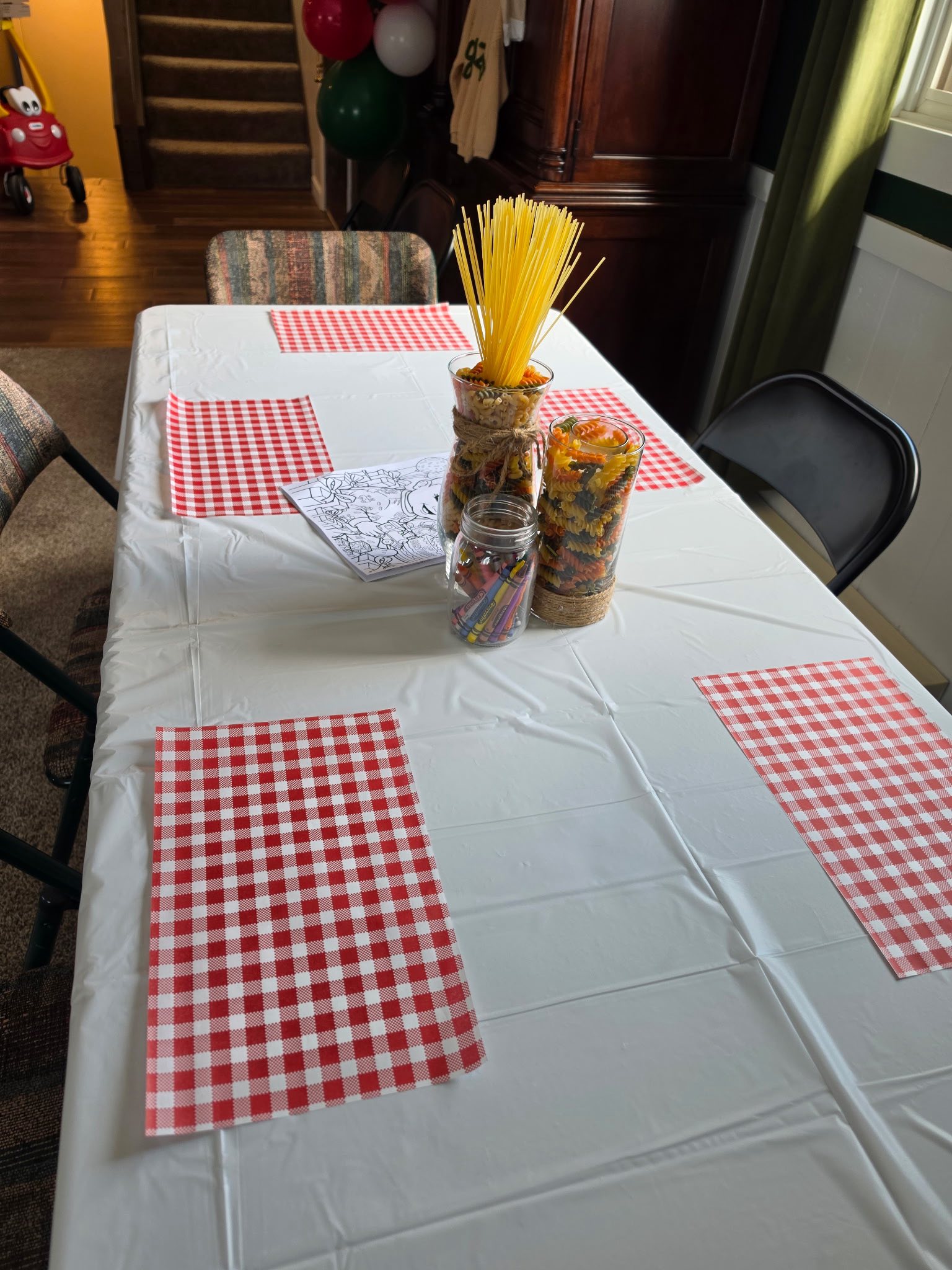 Dining table set for a pasta-themed party with red and white checkered placemats, pasta centerpieces, and coloring.