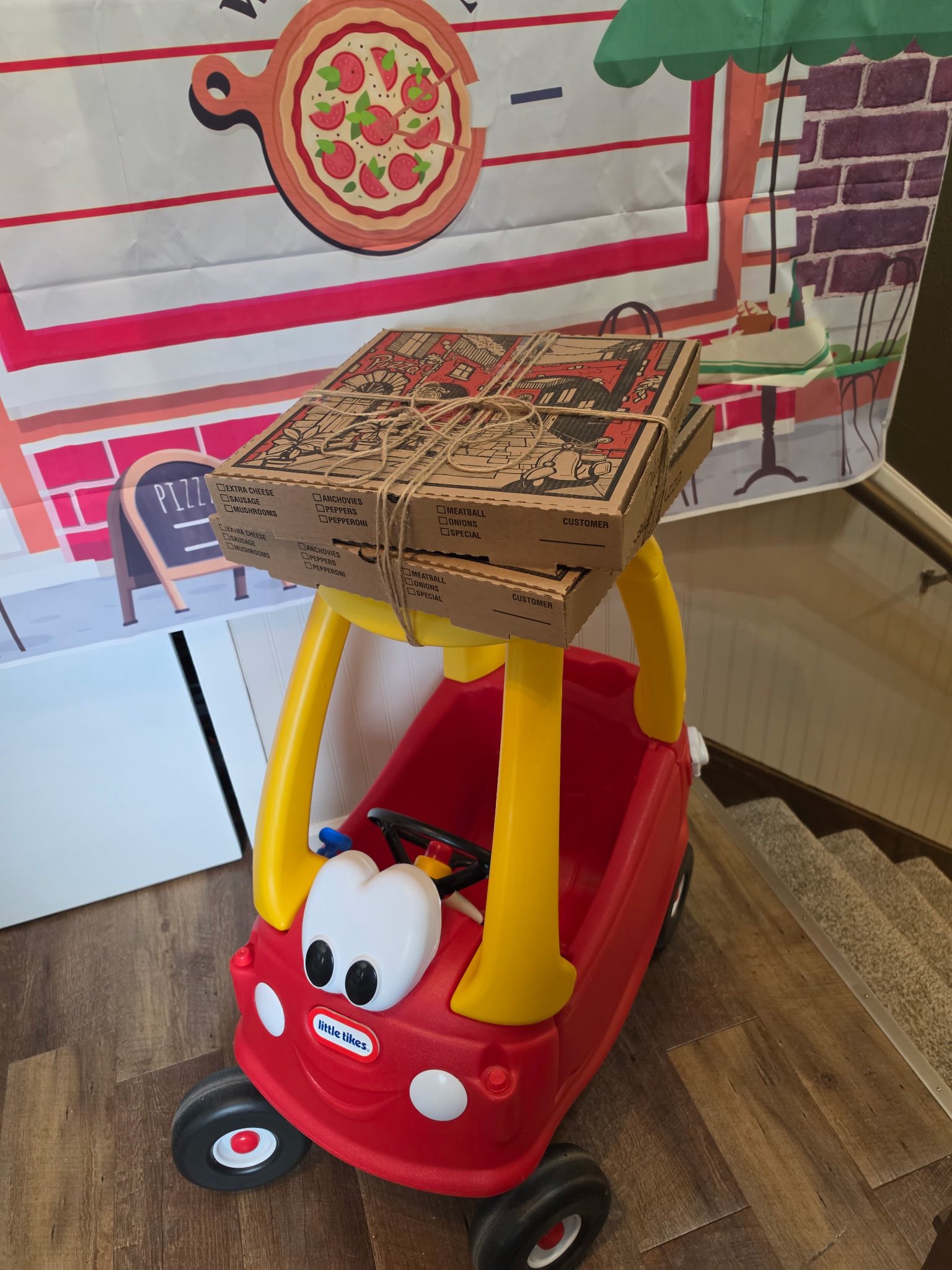 Two stacked pizza boxes tied with twine resting on the roof of a red and yellow Little Tikes toy car