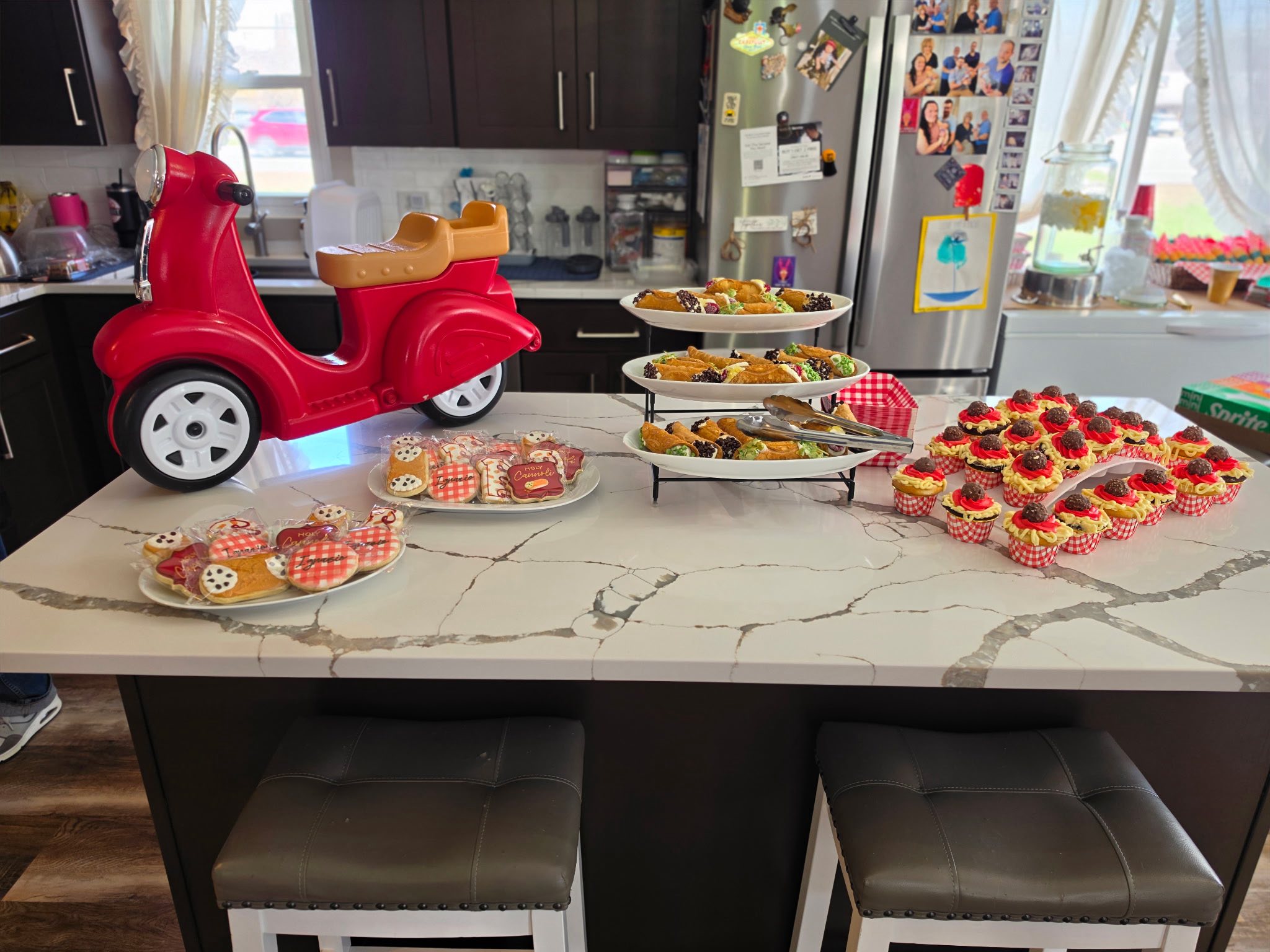 Kitchen counter spread featuring plates of iced cookies, cannoli on a tiered stand, and small cupcakes.