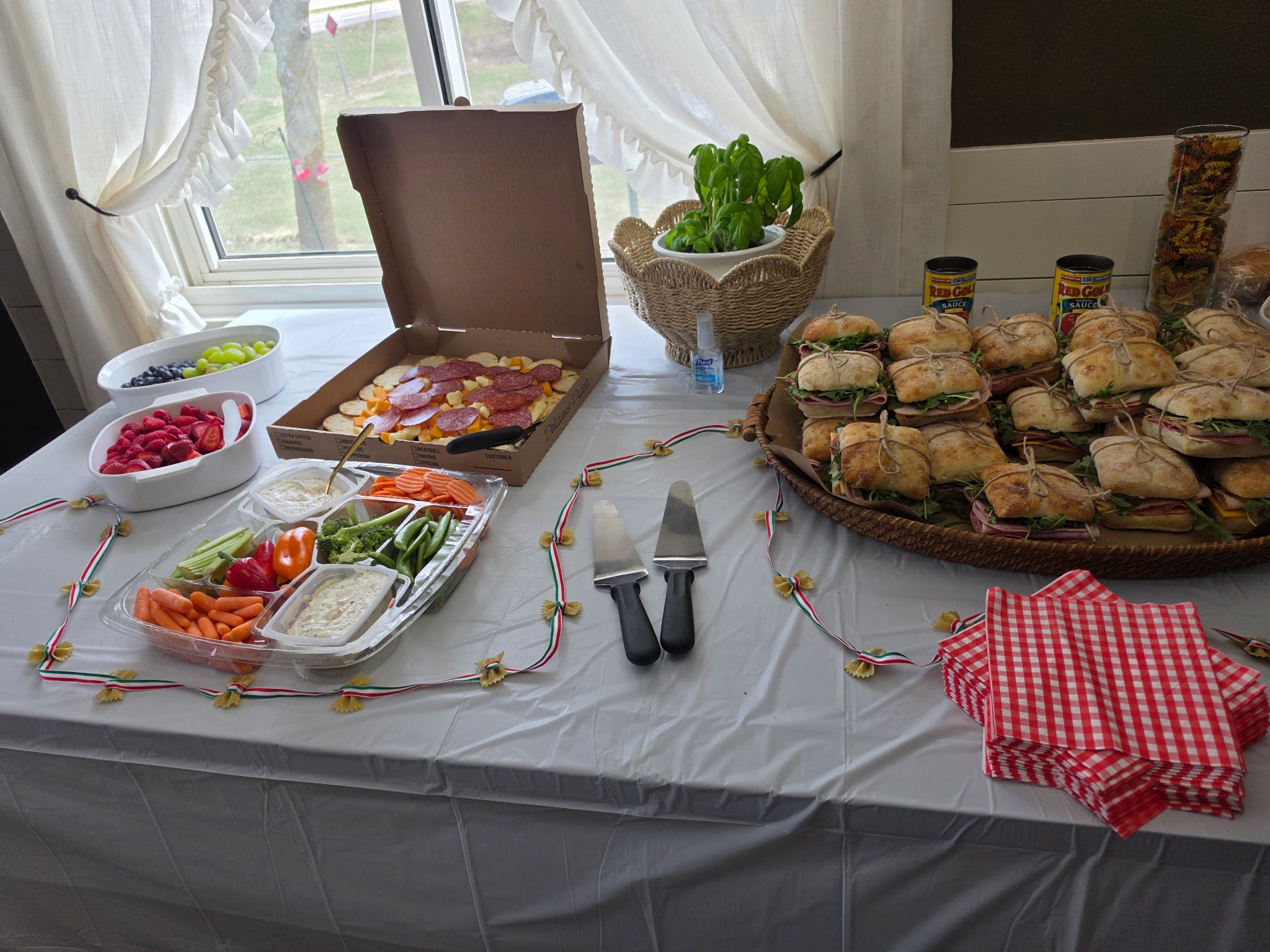 Table spread featuring a pizza box, charcuterie-style sandwiches, fresh vegetable platter, and fruit bowls.