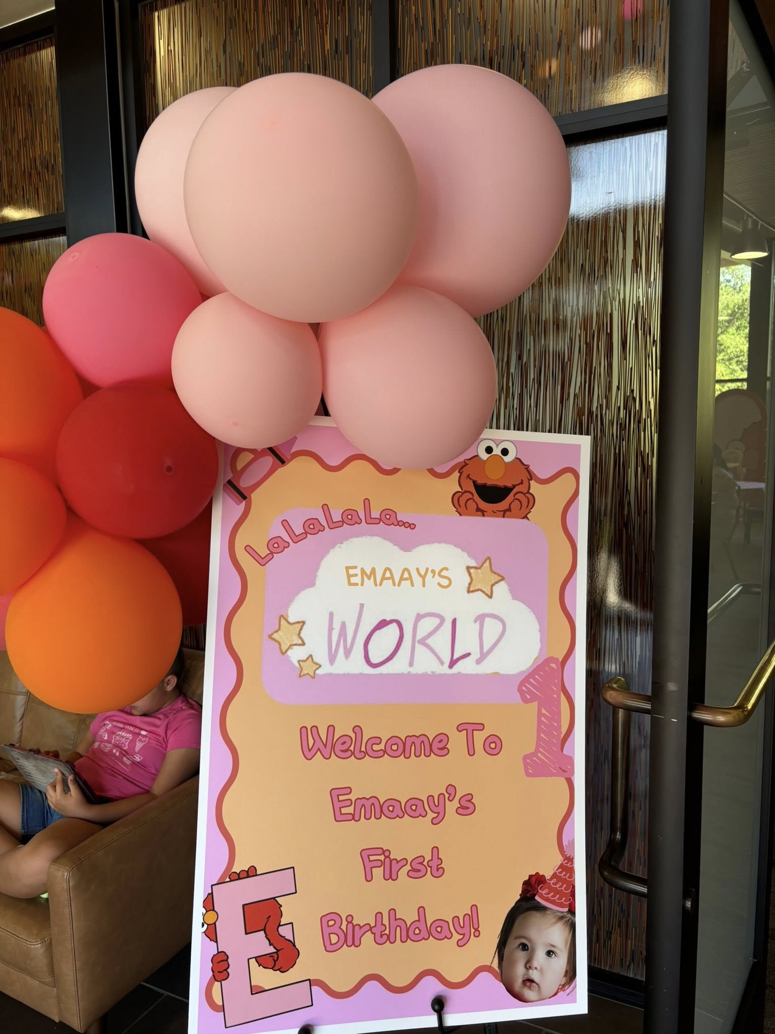 Elmo-themed first birthday welcome sign for Emaay decorated with pink and orange balloons.