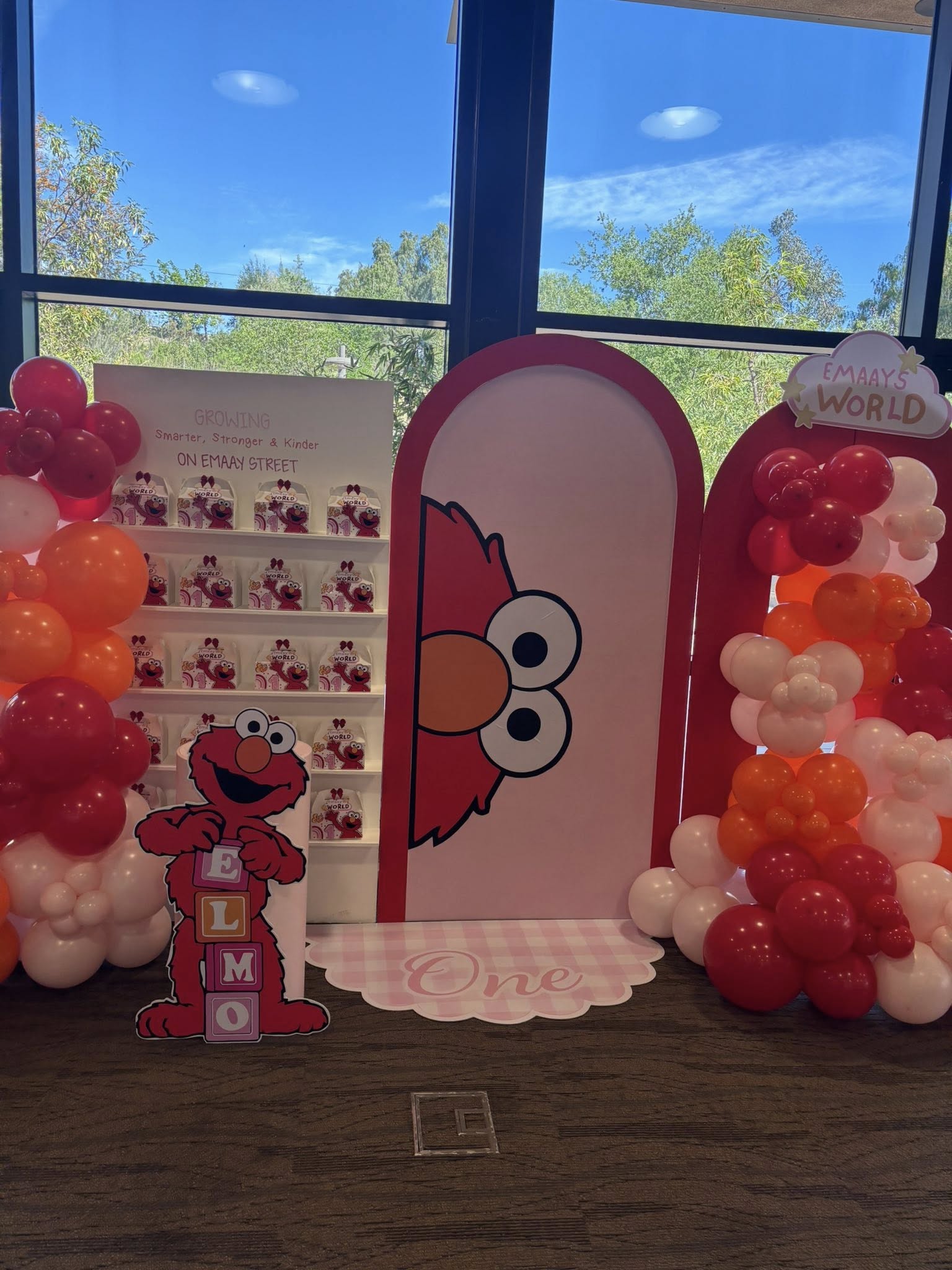 Sesame Street-themed Emaay's World first birthday display featuring Elmo props, balloon arches, and party favors.