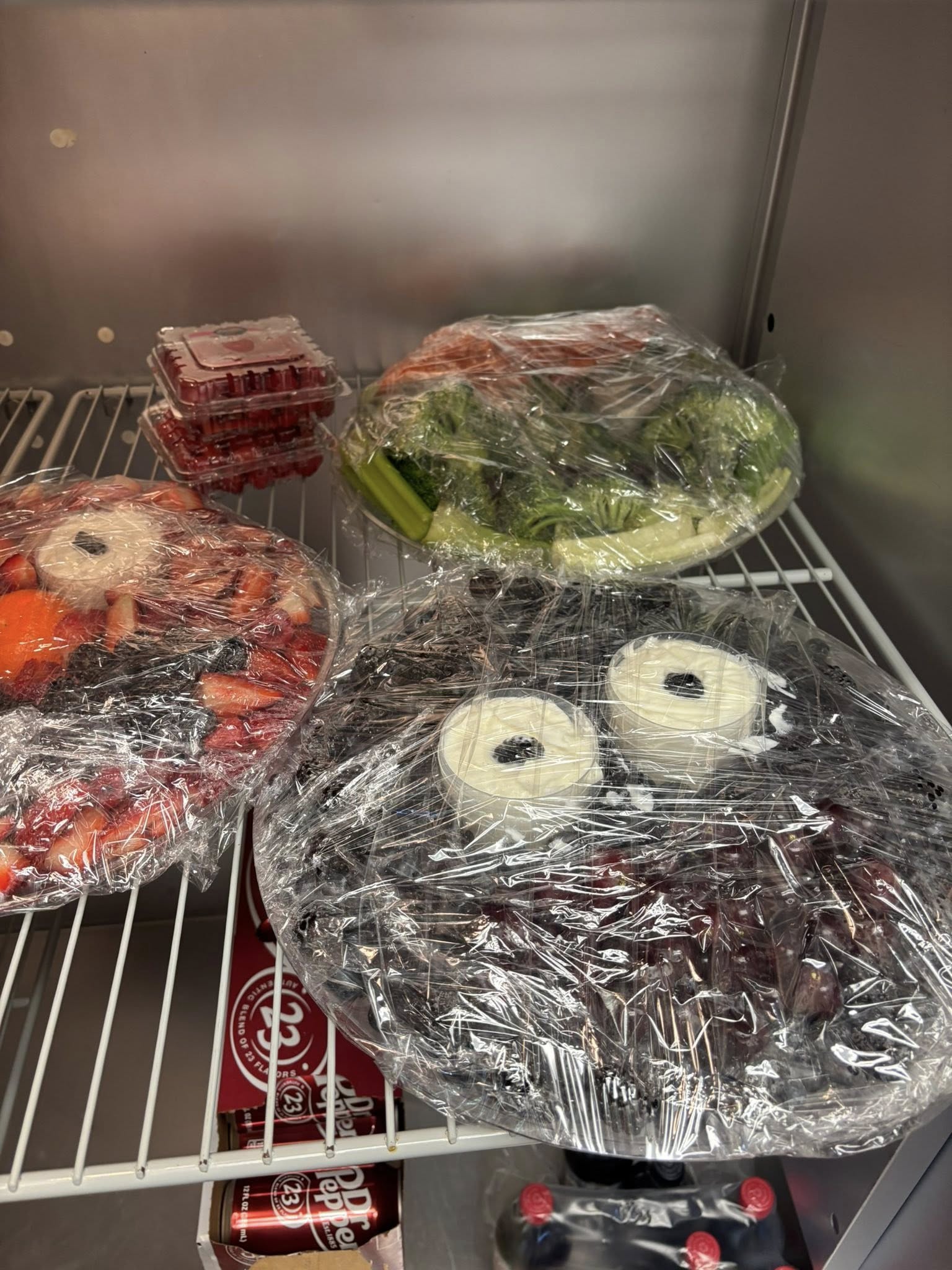 Plastic-wrapped fruit and vegetable platters with dip in a refrigerator shelf.