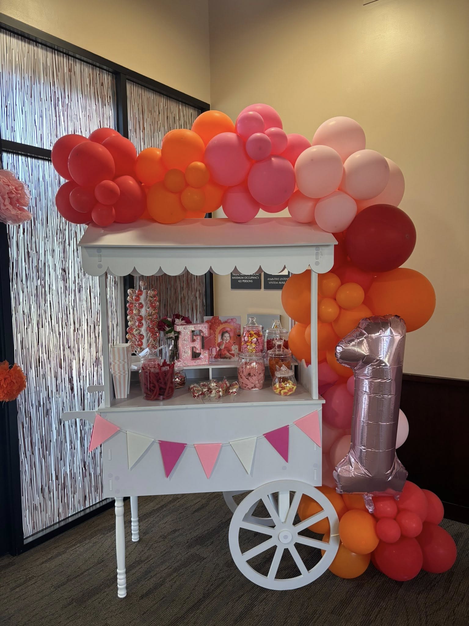White party treat cart decorated with a pink and orange balloon arch and a metallic number one balloon for a first birthday