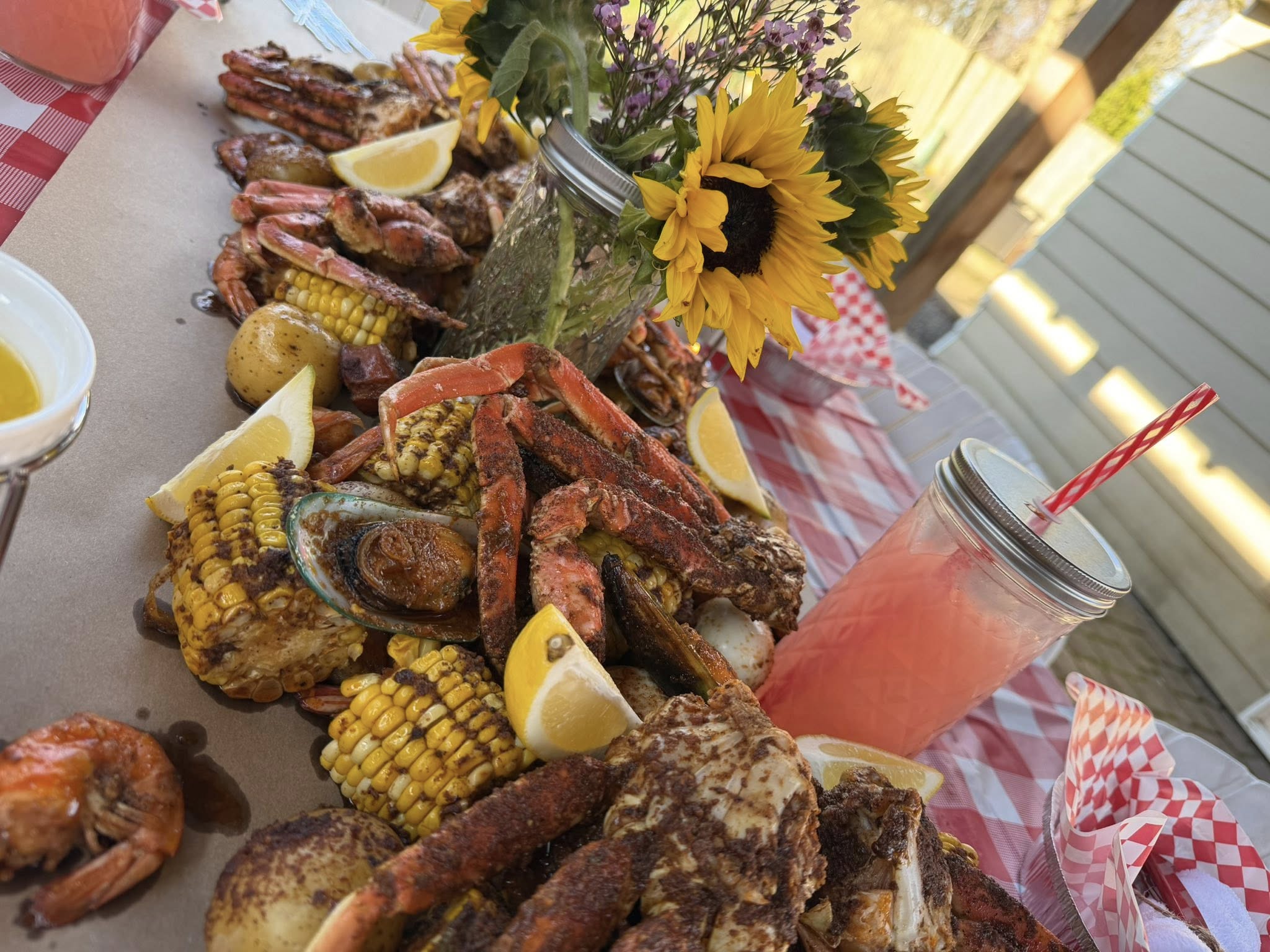 Seafood boil with crab legs, mussels, corn, and potatoes, styled with a sunflower centerpiece and pink drink.