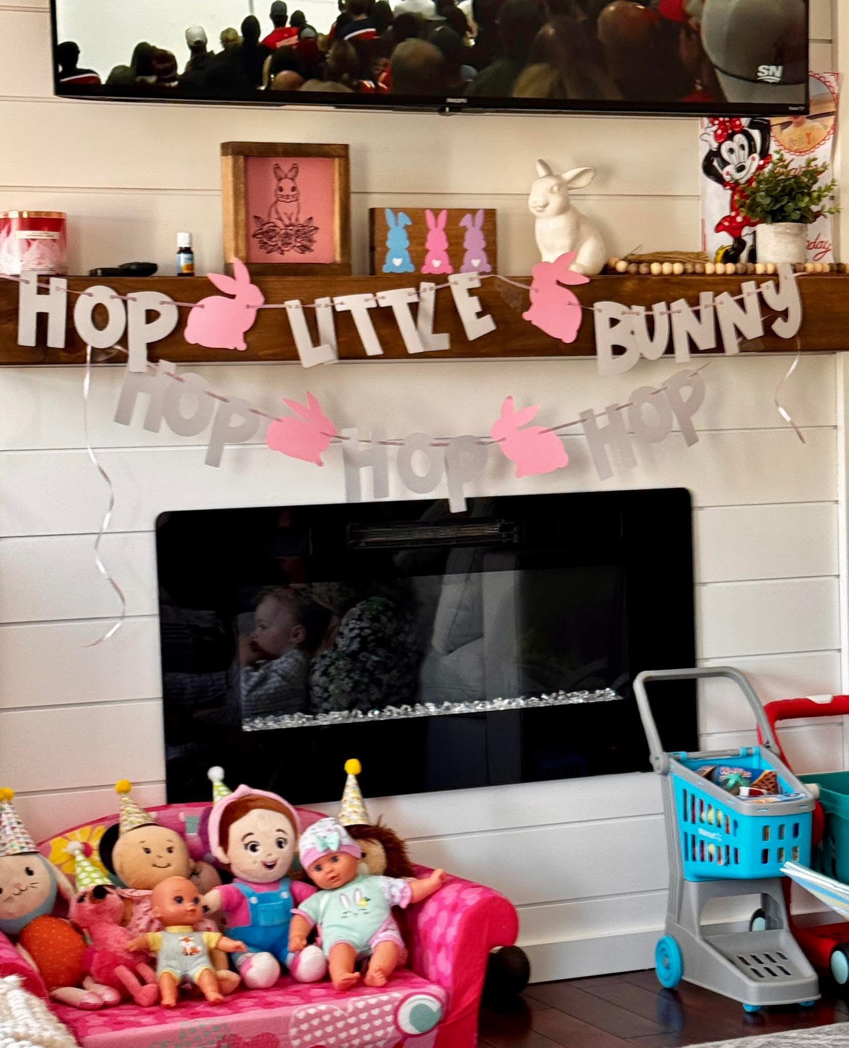 Easter-themed fireplace mantel decorated with a Hop Little Bunny garland, rabbit figures, and children's toys.