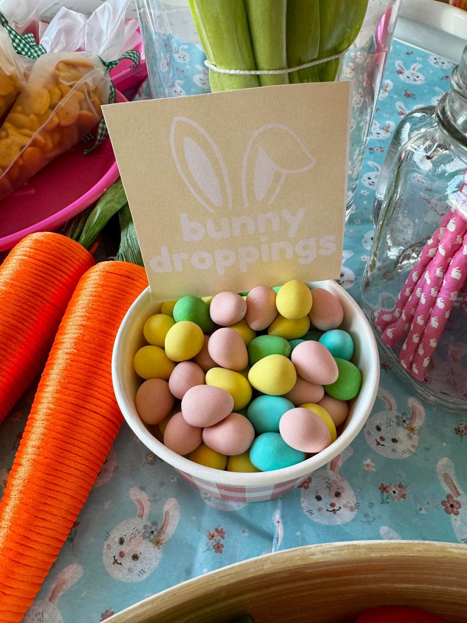 Bowl of pastel-colored chocolate eggs labeled bunny droppings on a table with Easter decorations