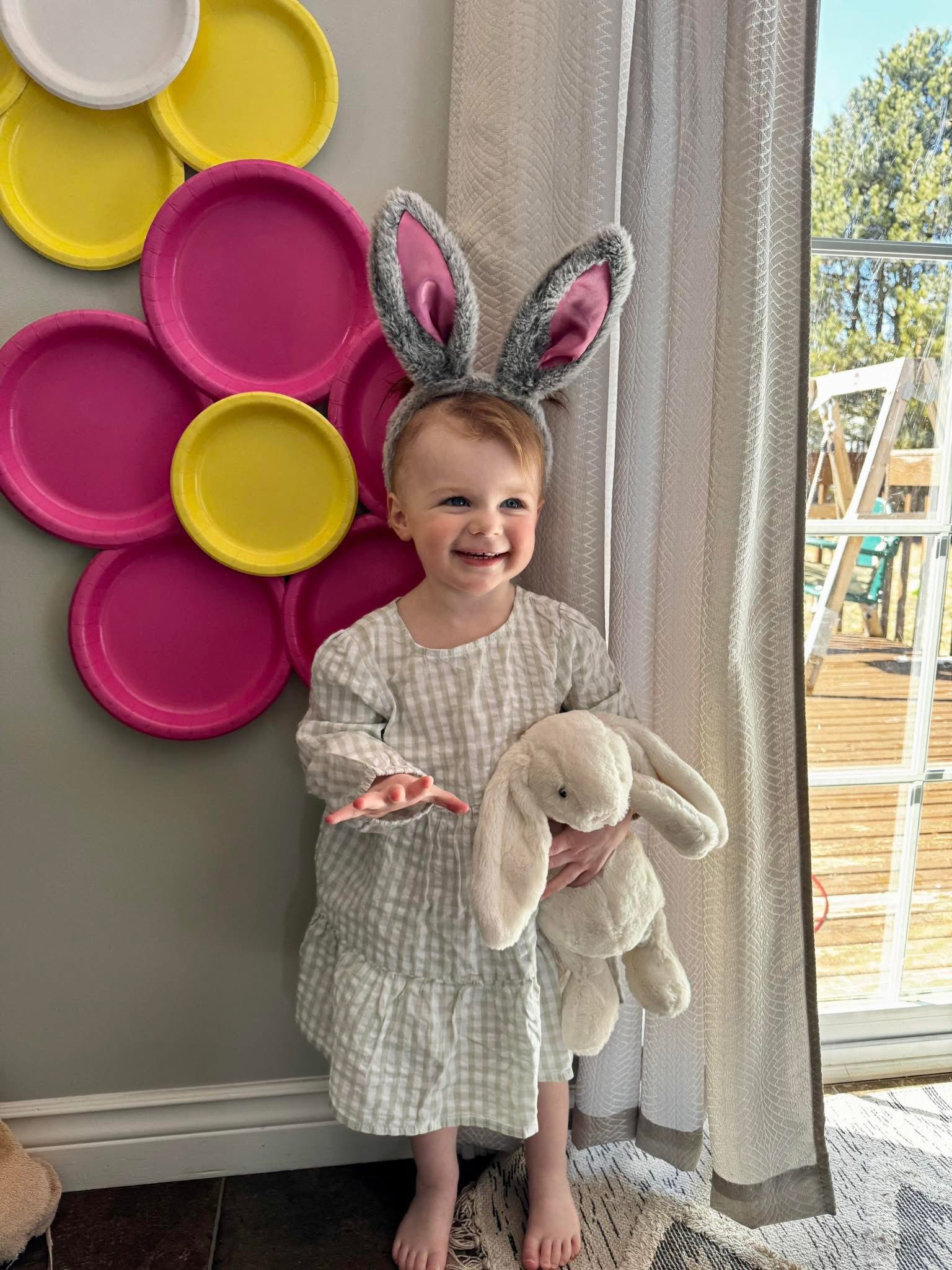 Toddler wearing fuzzy bunny ears and holding a white plush bunny rabbit, standing in front of a DIY paper plate flower.