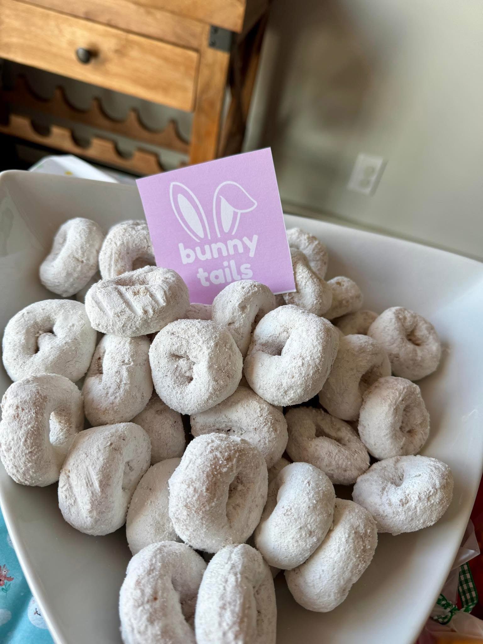Bowl of powdered sugar mini donuts labeled with a pink bunny tails sign for Easter.