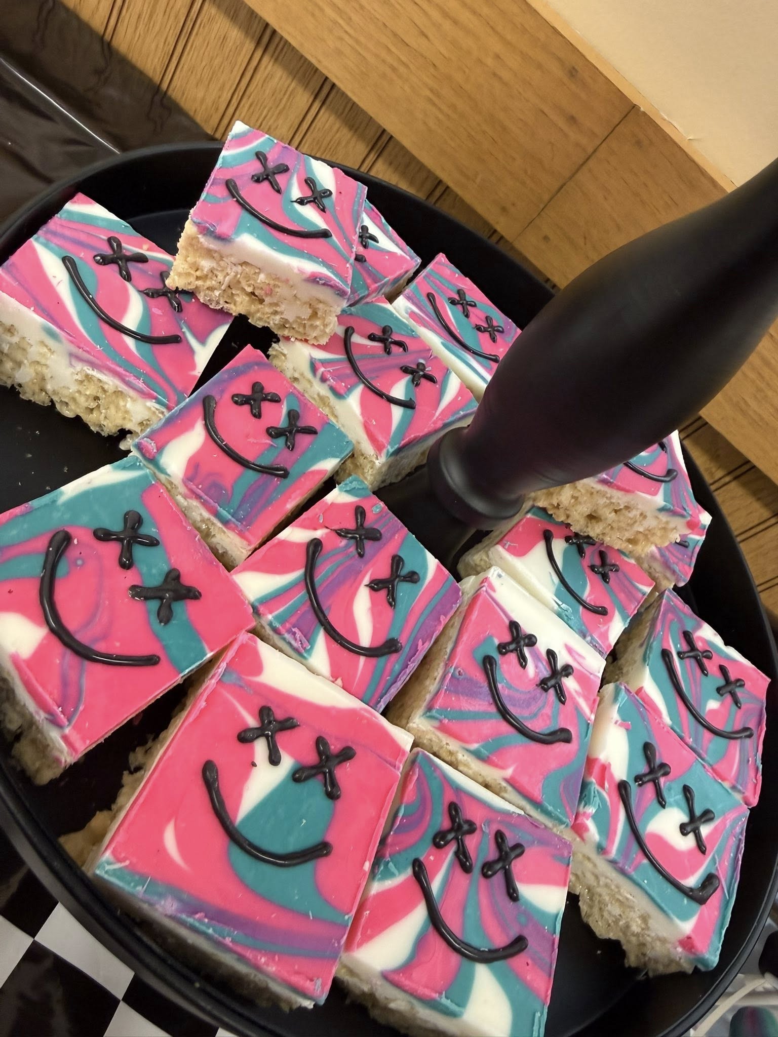 Square rice crispy treats with pink, blue, and white marbled icing and black smiley face piped designs on a tiered serving tray.