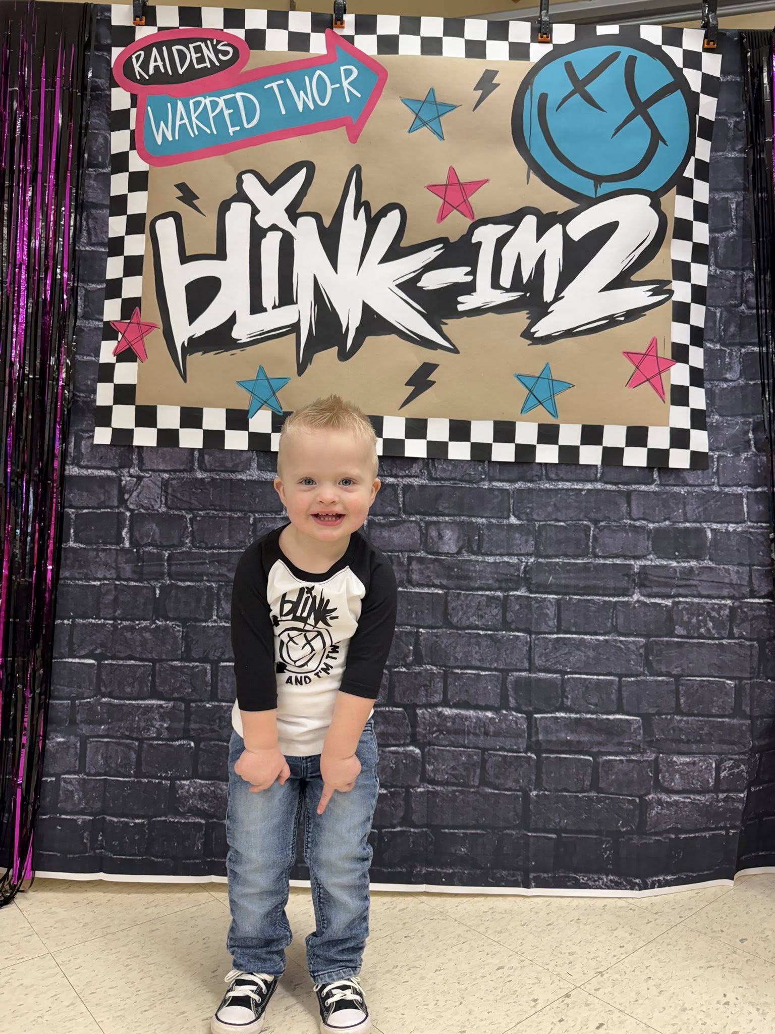 A smiling young boy wearing a Blink-182 themed baseball tee and jeans standing in front of a Blink-182 party backdrop.