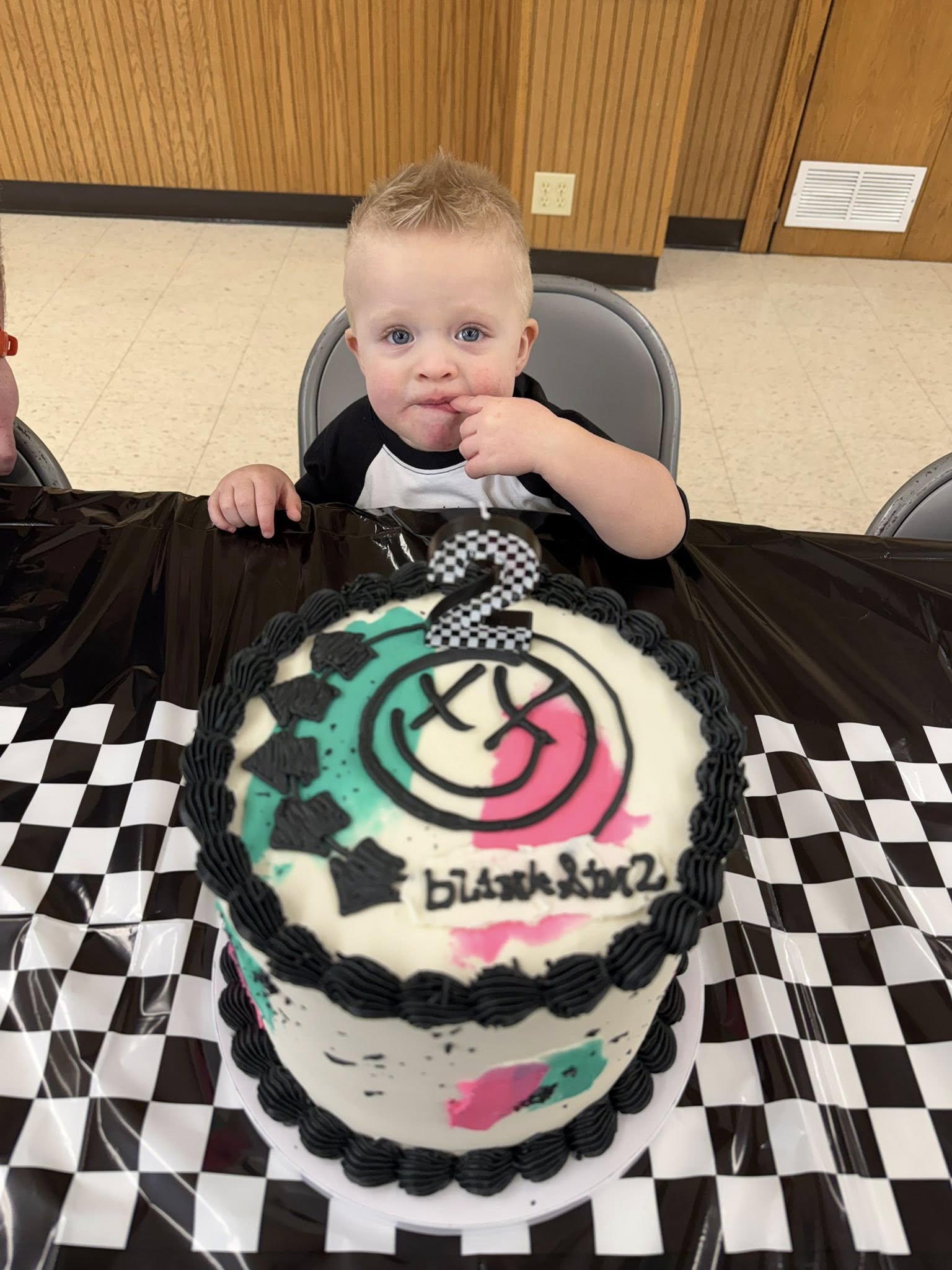 Toddler sitting at a table with a custom blink-182 themed birthday cake featuring a checkboard number two candle.