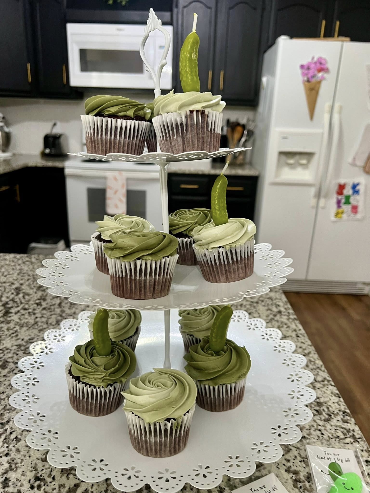 Cupcakes with green frosting topped with miniature pickles on a three-tier white dessert stand.