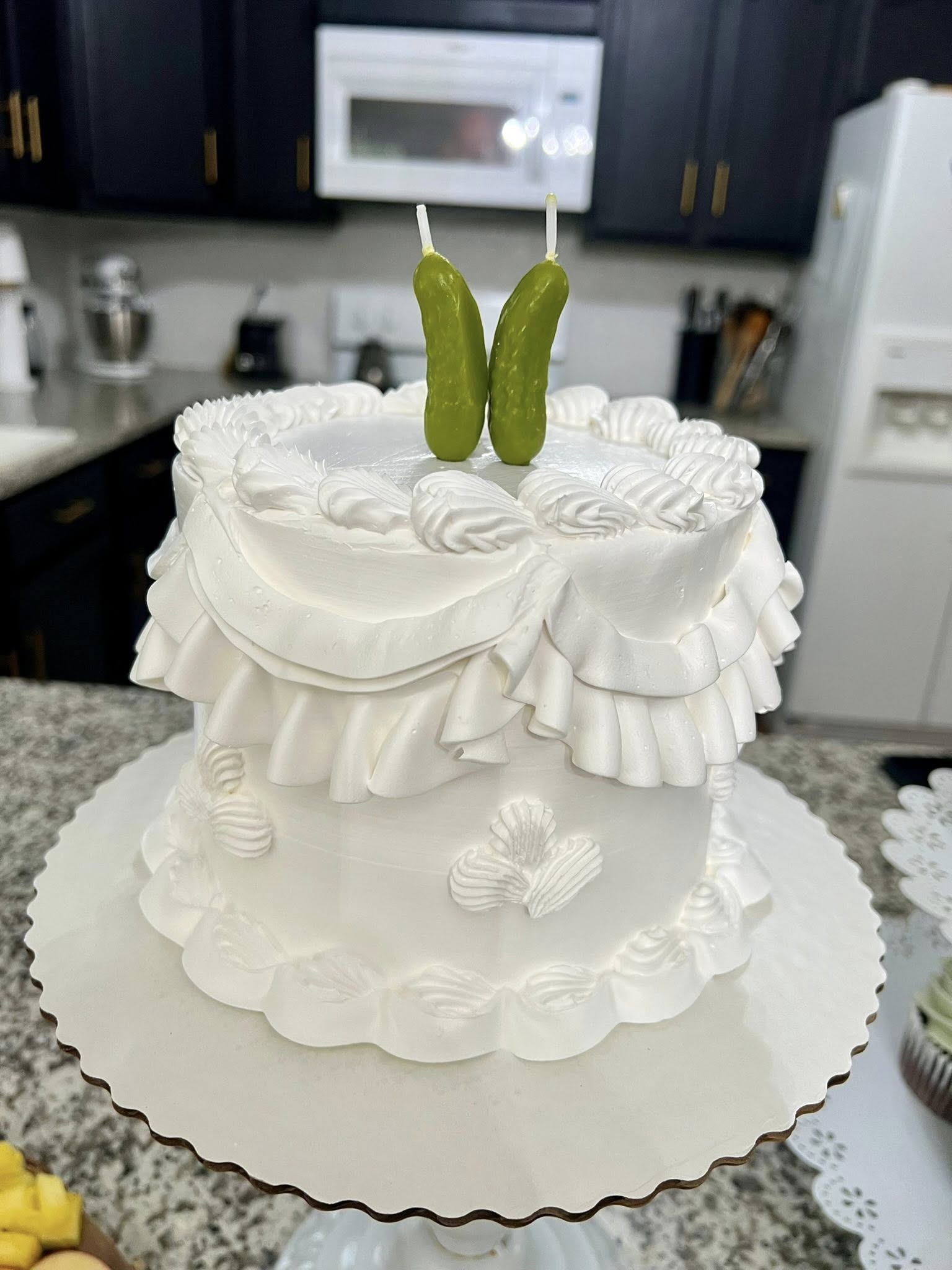 Round white cake with elegant buttercream ruffles and two green pickle candles on top.
